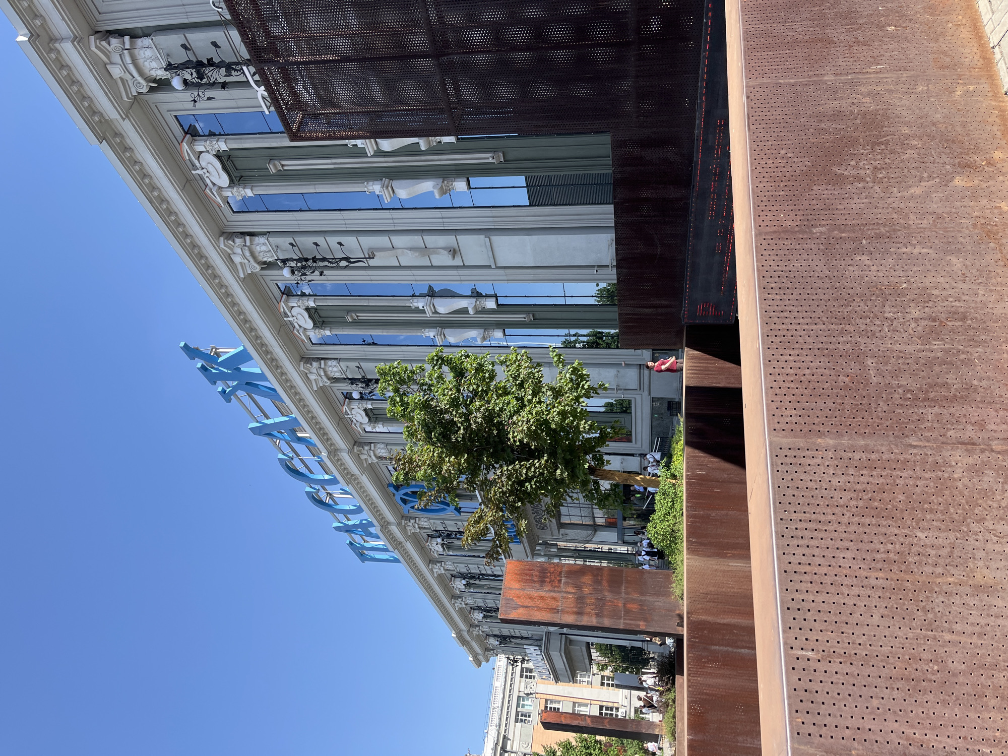 The image depicts a low-angle view of a building with classical architectural elements and modern additions. The building's facade is white with decorative features such as ornate moldings, columns, and classical ornaments. Above the building’s entrance, there is a blue structure with the letters "MAKEDO" arranged vertically. The windows reflect the sky and surrounding buildings.
In the foreground, there are large, rusted metal panels with a grid of perforations, partially obscuring the view of the building. These appear to be part of a modern art installation. Between the metal panels and the building, there's greenery including a small tree.
The background reveals additional buildings and what appears to be pedestrian traffic, indicating an urban environment.
Given the "MAKEDO" inscription, and the architectural style of the building in question, this appears to be the Makarov Bridge and surrounding area in Yekaterinburg, Russia. The rusty perforated metal panels may be a part of the installation of the pedestrian alley and museum in the city.
