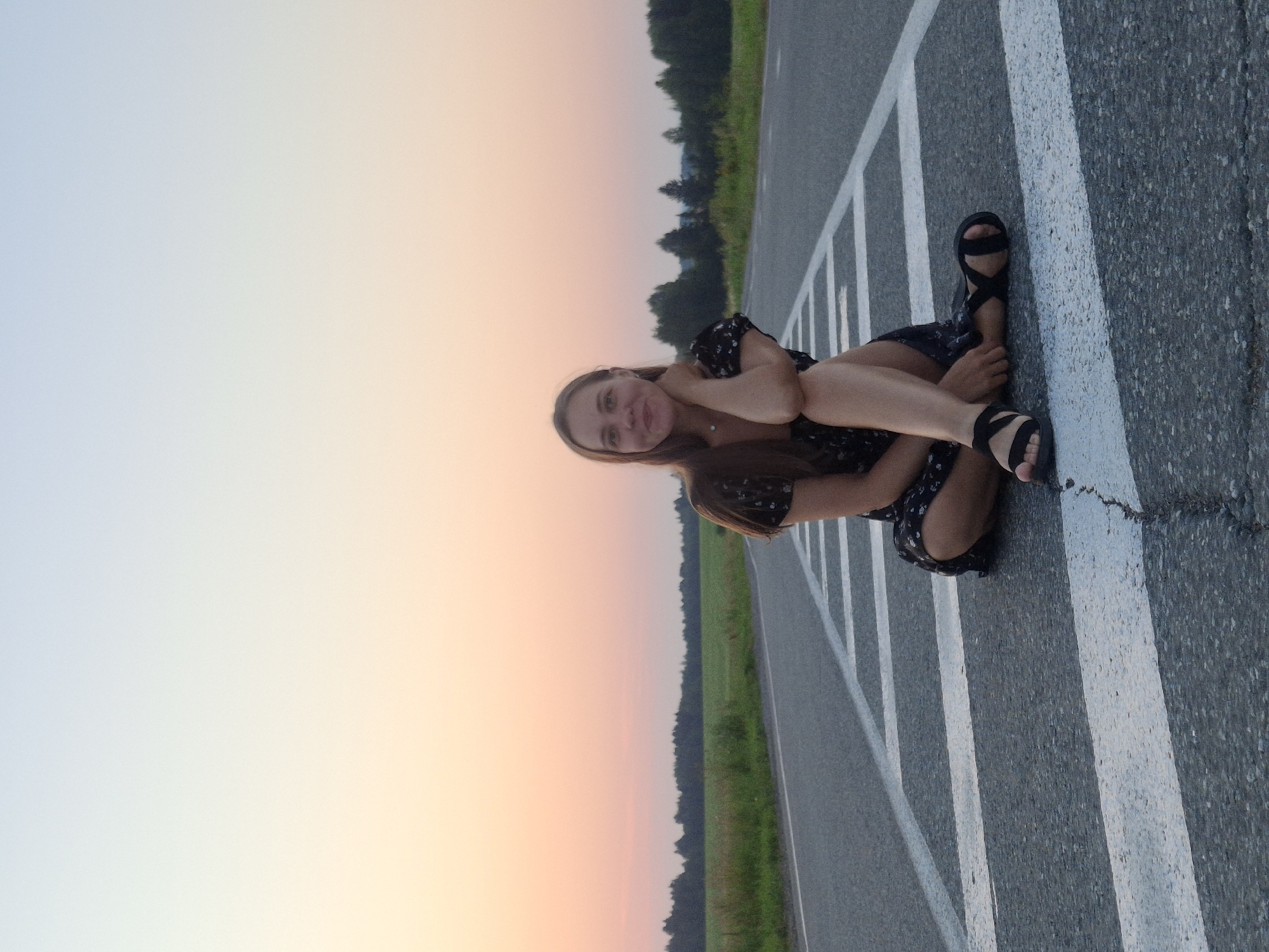 A young woman with long brown hair is sitting cross-legged on an asphalt road. She is wearing a dark, floral-patterned dress and black sandals. Her right hand is resting on her chin, and she is looking towards the camera with a slight smile. The road has white markings. In the background, there is a green field and a line of trees. The sky is a gradient of pale blue and pink, suggesting it is either sunrise or sunset. The image appears to be taken outdoors, possibly in a rural area outside the city of Perevoznoe, Russia.