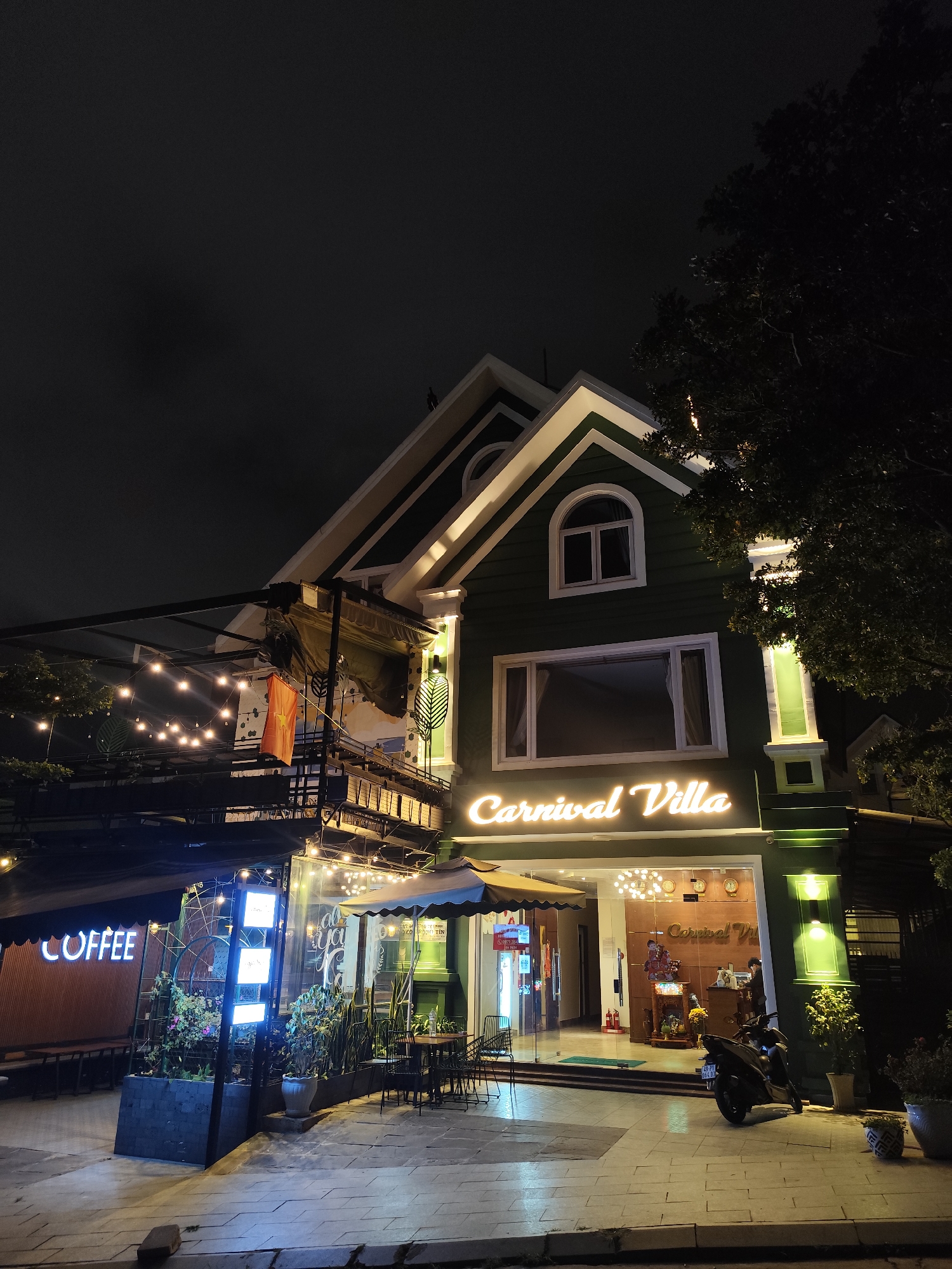 The photo depicts a night scene in Da Lat, Vietnam, featuring the Carnival Villa. The building is two stories, green, and white, with a prominent "Carnival Villa" sign lit in neon. A coffee shop is located to the left of the villa, with "COFFEE" spelled out in neon lights. Tables and chairs are set up outside. A motorcycle is parked in front of the Villa. The sky is dark. Decorative string lights are suspended. A Vietnamese flag is visible above the coffee shop area. Some people are at the front desk inside the villa. Vegetation (trees and potted plants) is present. The pavement is composed of light-colored paving stones.