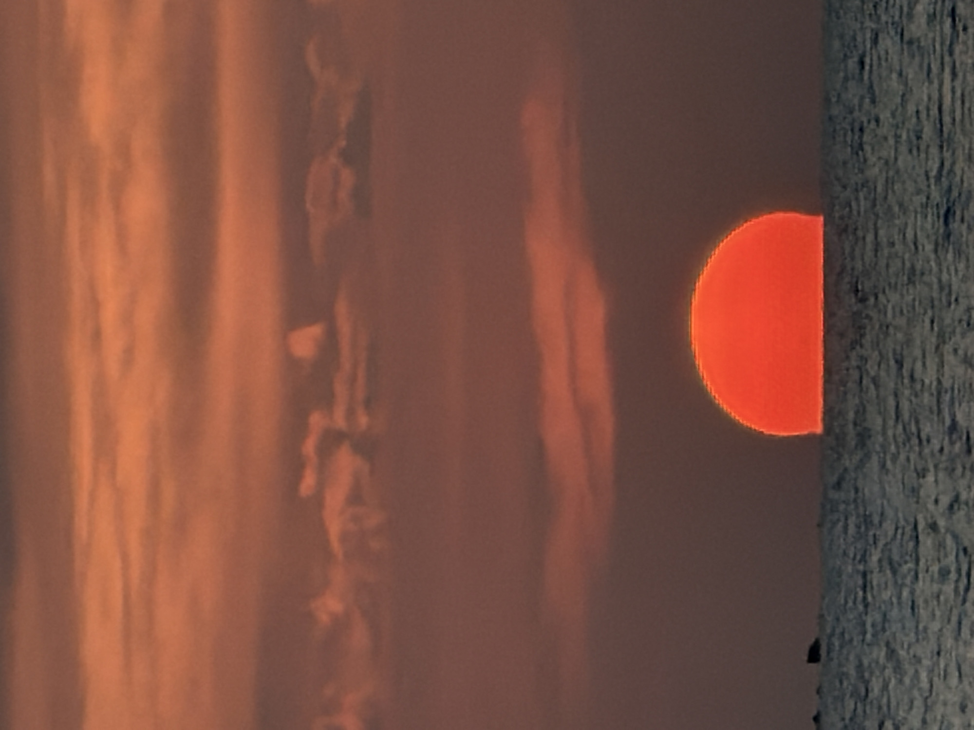 An extreme close-up shot captures the lower half of a brilliant orange sun sinking below the horizon of the ocean at sunset. The sun is a perfect semi-circle, emitting a warm glow that washes over the scene. The water is a deep blue, reflecting the sky's colors and creating a textured surface with subtle ripples. Wispy, light orange clouds are scattered across the sky, adding depth and dynamism to the atmospheric backdrop. In the distance, a dark, indistinct silhouette of land or a boat is visible on the left side of the frame, providing a sense of scale and context. The overall mood is serene and tranquil, evoking the peaceful beauty of a coastal sunset.