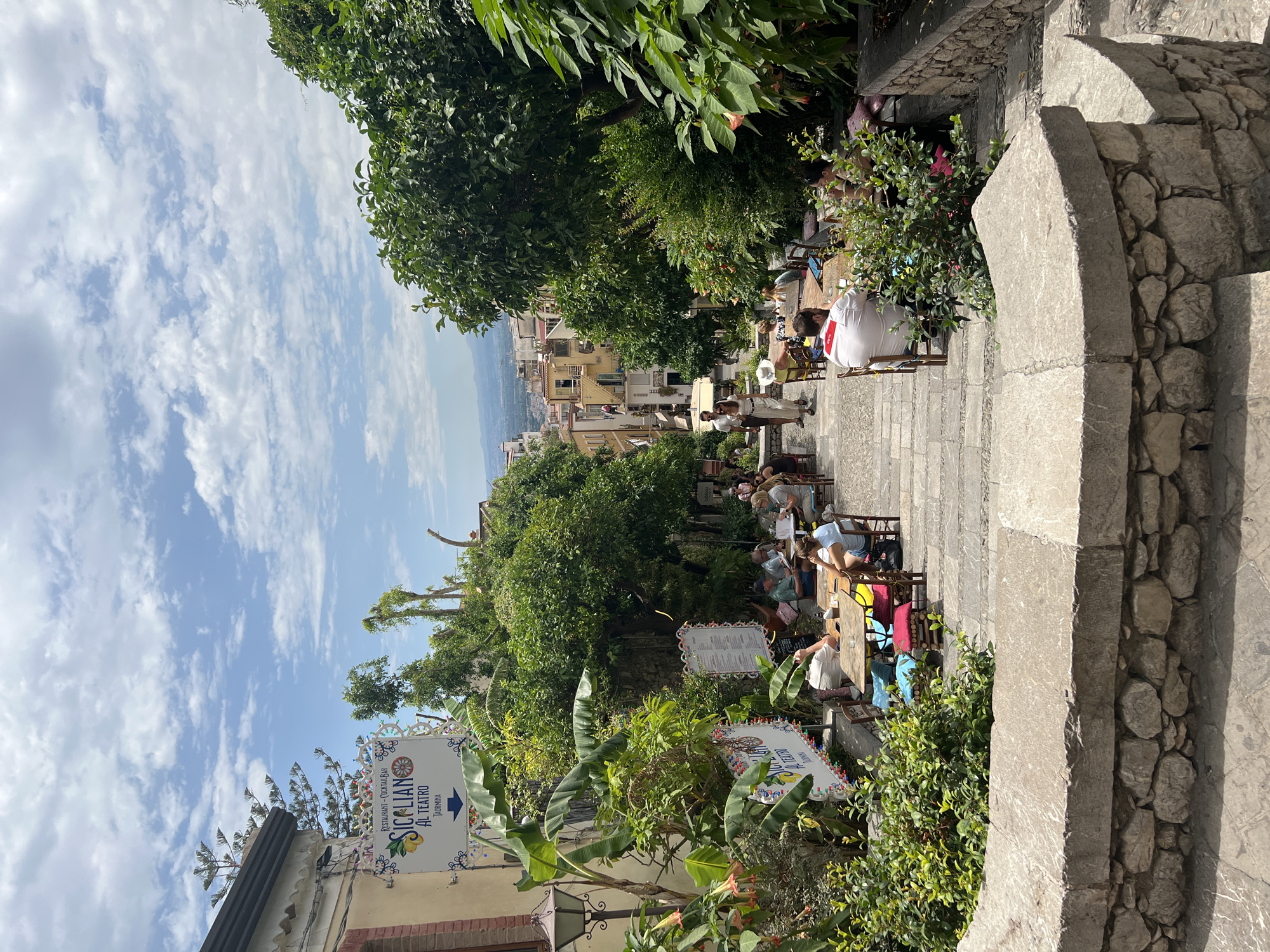 The image shows a steep, stone-paved stairway in the city of Taormina, Italy, lined with potted plants, trees, and outdoor seating areas for cafes and restaurants. Several people are seated at tables, enjoying meals and drinks. A sign for "SICILIAN AL TEATRO" is visible on the left side of the stairway, indicating a restaurant or cafe. The stairway leads down a street with buildings and a panoramic view of the town and the distant landscape. The sky is partly cloudy, with a bright blue expanse.