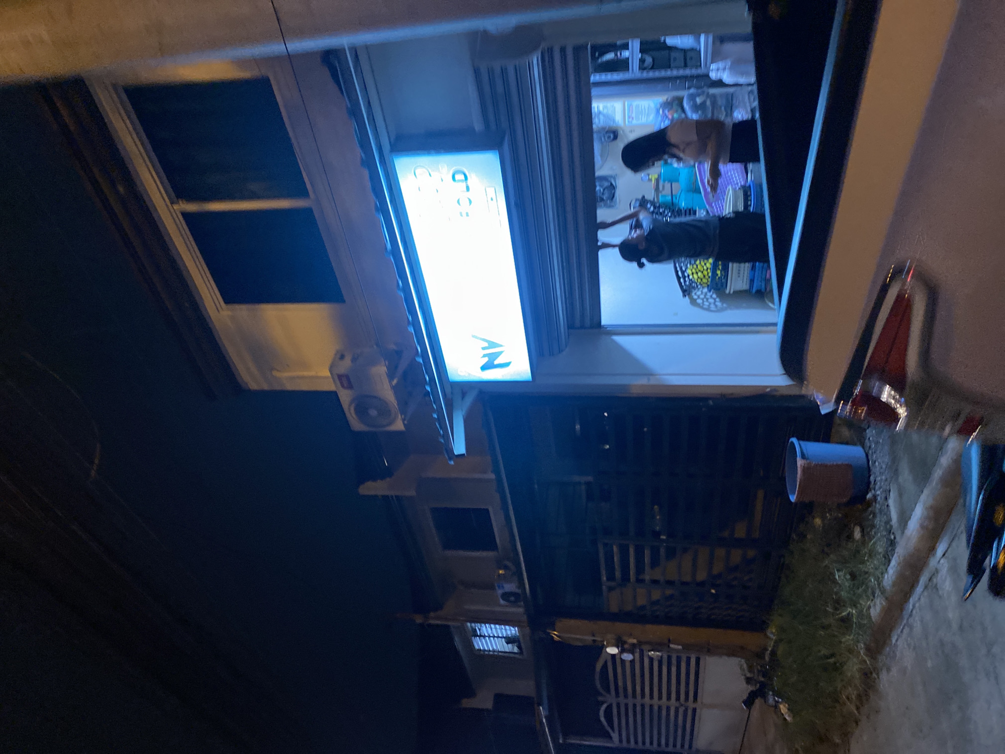 This media file captures a night scene outside a building, likely a commercial establishment, in Davao City, Philippines. The overall image is dark, illuminated primarily by artificial light sources.
The central subject is a shopfront with a large glass window, revealing its brightly lit interior. Above the shop entrance, a rectangular light box sign is prominent. When viewed correctly, the sign displays the letters "AN" on its left side and the word "FOLD" on its right, possibly indicating a laundry or dry-cleaning service. Two small circular logos are visible above the word "FOLD." An air conditioning unit is mounted on the wall below the sign.
Inside the shop, two individuals are visible through the glass. One person, with long dark hair, stands further back near colorful baskets. The second person, closer to the front, with dark hair tied back, is leaning over a basket containing yellow and white patterned items, possibly laundry. Both appear to be engaged in work, likely sorting or processing clothes. The shop interior is functional, with various baskets and shelving units visible.
To the left of the shopfront, the building continues with windows that appear dark, suggesting unlit rooms or a residential section above. Further left and below, a metal gate is visible, possibly another entrance to the building or an adjacent property.
On the ground in front of the shop, a blue bucket with a brown rim is situated near a patch of grass and a concrete pavement. Partially visible on the right side of the frame are parts of what seem to be parked vehicles, indicated by a red taillight and dark bodywork. The street appears quiet, typical of late evening or night. The scene depicts a mundane, operational business environment during non-daylight hours.