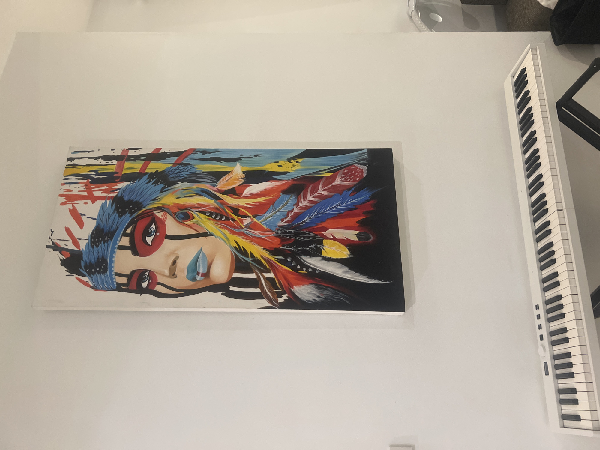 A vertical, medium shot captures a painting of a woman with Native American-inspired headdress and face paint, displayed on a white wall in what appears to be a domestic interior in Ko Samui District, Thailand. The painting, rendered in a vibrant, abstract style, features the woman's face with striking blue lips and eyes, framed by a blue headdress adorned with multicolored feathers. To the left of the painting, a white electronic keyboard with black and white keys rests on a black stand, its surface illuminated by ambient light. The wall is painted a neutral white, providing a clean backdrop for the artwork and musical instrument. The overall scene suggests a personal living space, possibly a studio or music room.