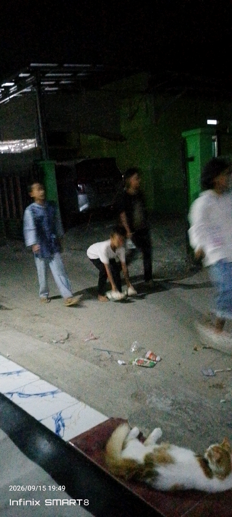 In Serang, Indonesia, this nighttime scene captures a street with several young individuals. One person, wearing a blue shirt and light-colored pants, stands to the left. Another, in a white t-shirt and dark pants, is crouched down in the center, appearing to interact with something on the ground. To the right, partially visible, another person in a dark shirt is standing. Further to the right, a person in a white shirt is blurred, suggesting movement. Scattered on the ground are various pieces of litter, including plastic bottles and wrappers. In the foreground, a ginger and white cat is lying on its back on a reddish-brown surface. The environment includes dark buildings and a green structure in the background. A timestamp "2026/09/15 19:49" and "Infinix SMART8" are overlaid on the bottom left of the image.