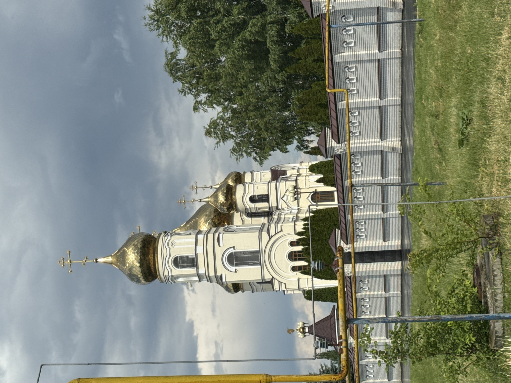A golden-domed cathedral stands tall against a cloudy sky in Alekseyevka, Russia. The cathedral's facade is a pale cream color with white trim, featuring multiple stories and windows. Several smaller golden domes, each topped with a cross, surround the main dome. A white brick wall with decorative arches and a dark metal gate runs horizontally in the foreground, separating the cathedral from a grassy area. Yellow pipes are visible running along the top of the wall and a prominent yellow pole stands to the left of the frame. Green trees and bushes are scattered throughout the scene, with a dense line of cypress trees directly behind the wall and a large deciduous tree to the right of the cathedral. The ground in front of the wall is covered in green grass.