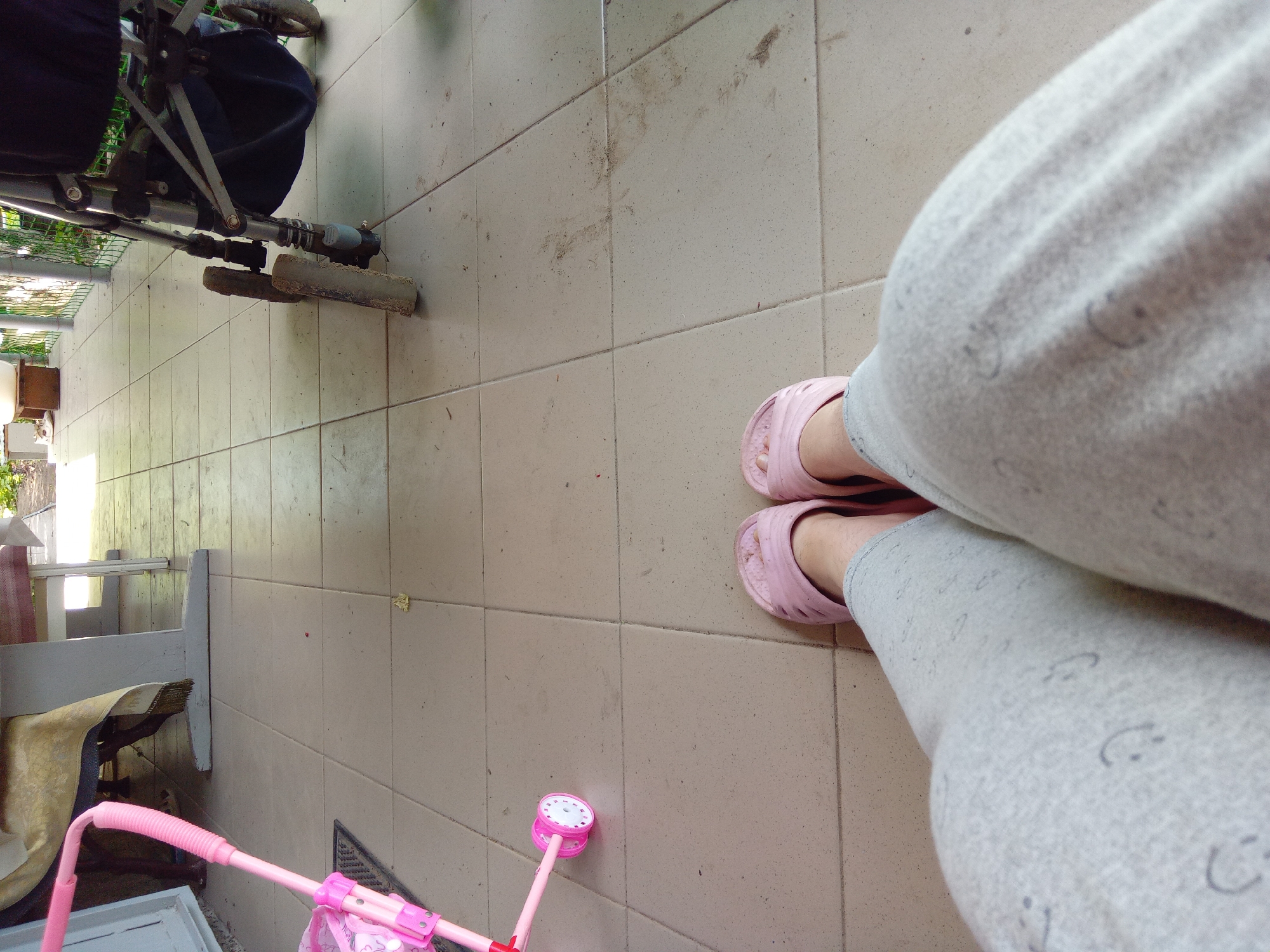 The image depicts a tiled porch with two pink rubber flip-flops on the feet of an individual wearing gray sweatpants with a subtle smiley face pattern. To the left, a pink toy stroller with white wheels is visible, positioned on a dark doormat. In the background, a portion of a wheelchair with a black seat is visible. Further back, a table covered with a patterned cloth is partially seen, with the legs of chairs underneath. The porch appears to be outdoors, with sunlight casting shadows on the tiled floor. The scene is set on a porch in Kitskany, Moldova.