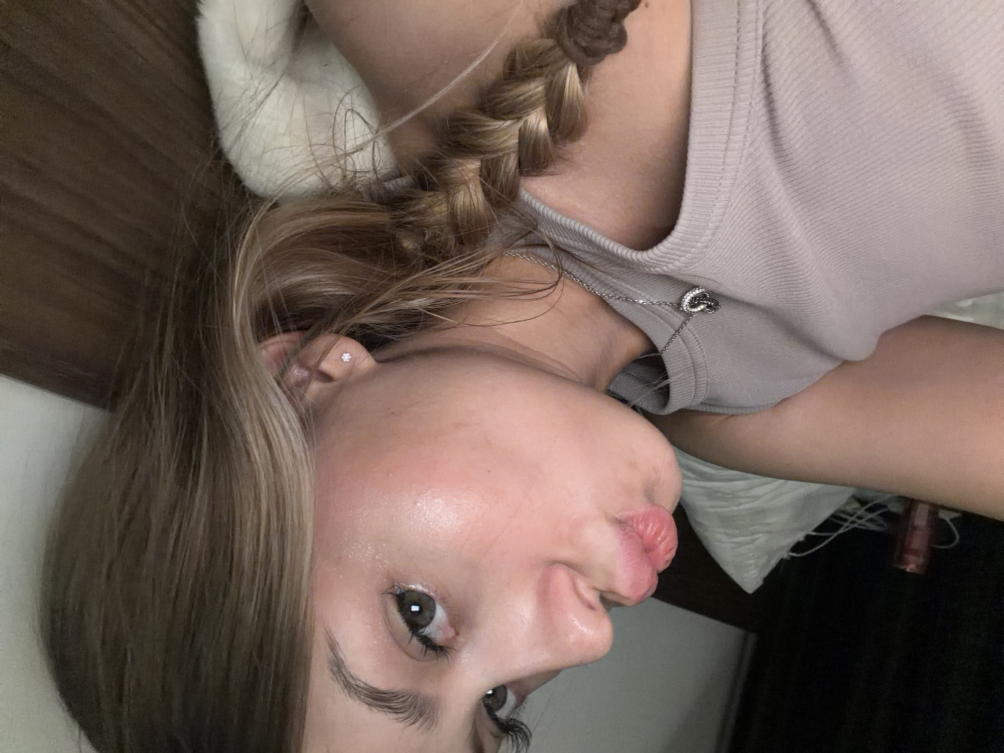 The image shows a close-up, rotated view of a young woman. She has blonde hair styled in a braid, a diamond stud earring, and is wearing a light-colored, ribbed tank top with a delicate silver necklace that has a heart pendant. Her face is turned, and she is making a puckering expression with her lips. The lighting suggests an indoor setting, possibly at night or in a dimly lit room. The background is blurred but hints at a domestic environment with textures that could be bedding or furniture. There is no visible text in the image.