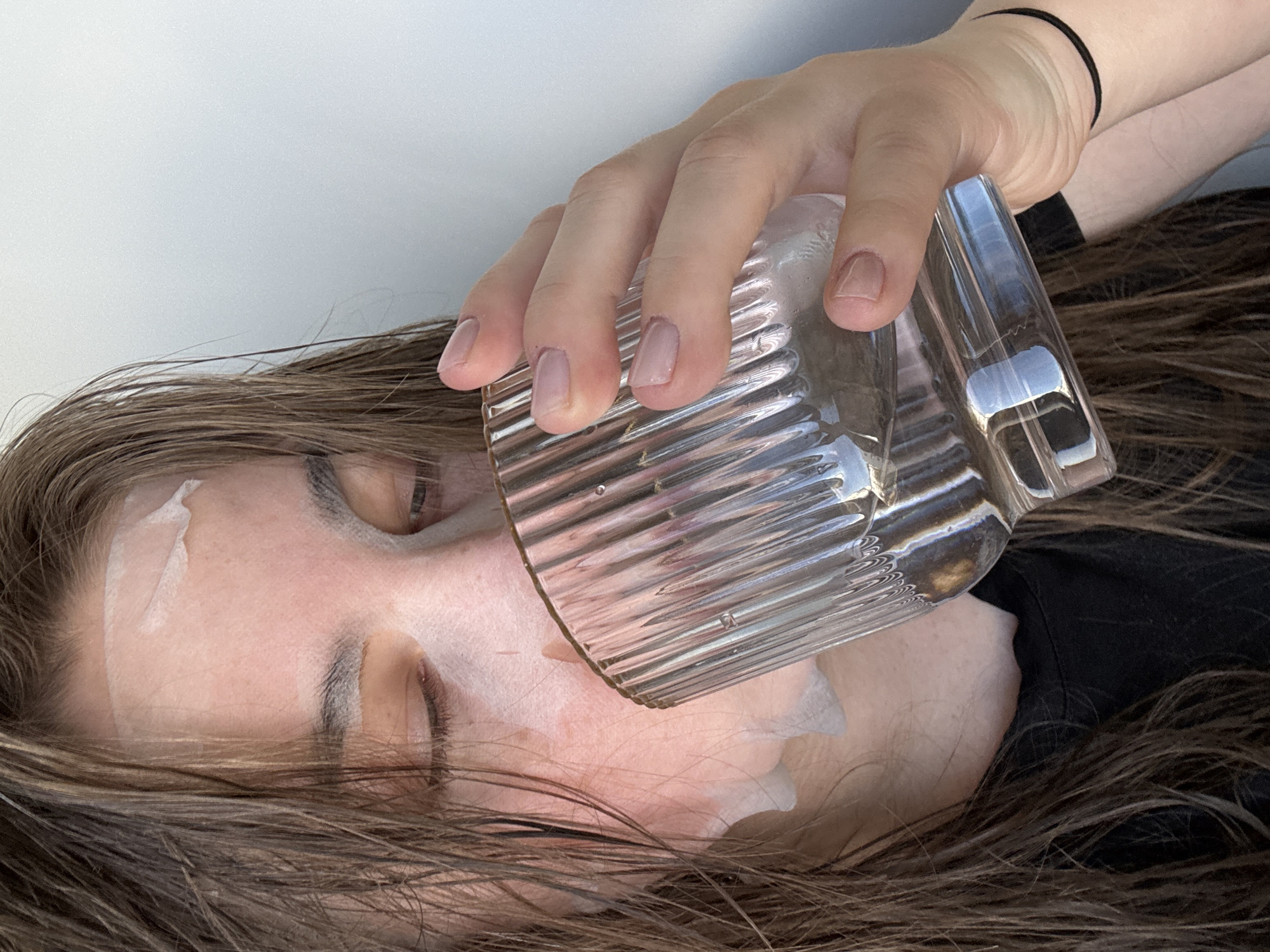 A close-up, rotated shot depicts a person, likely female, lying down with their eyes closed. Their hand is holding a clear, textured glass and tilting it towards their face. A thick, white, creamy substance, resembling a face mask or cosmetic product, is visibly applied to their forehead, cheek, and around the mouth. The person has long, brown hair spread around their head. They are wearing a dark, possibly black, garment, and a black elastic band is present on their wrist. The background is a plain, light-colored surface, suggesting an indoor setting with bright, diffuse lighting. No specific location cues are visible within the frame. There is no visible text. The activity appears to be the application or interaction with a face mask or similar product, possibly involving the glass. The scene is set in Chaikovsky, Russia.