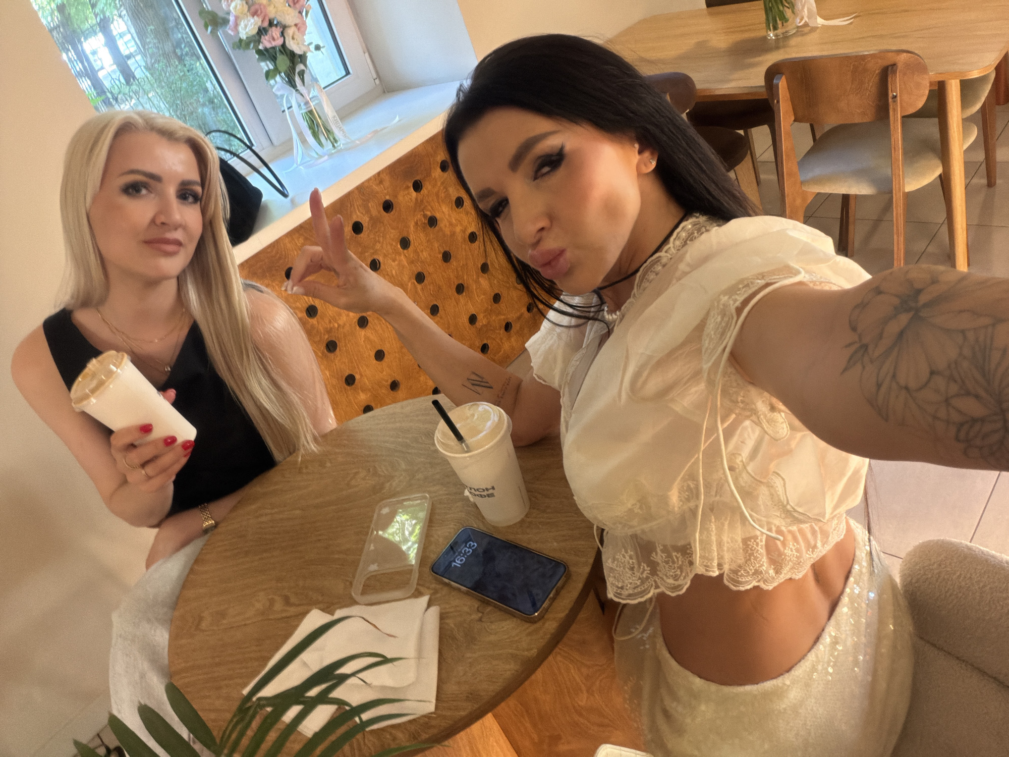 Two women are seated at a round, wooden table in what appears to be a cafe or restaurant. The woman on the left has blonde hair and is wearing a black top. She is holding a white cup with a straw in her right hand. The woman on the right has dark hair and is wearing a white, lacy top and a sequined skirt. She is making a peace sign with her left hand and pouting her lips. On the table, there are two phones, one with its screen displaying "16:33", a clear phone case, a white paper cup with a straw, and a stack of white napkins. A potted plant with green leaves is visible in the foreground. In the background, a window looks out onto trees, and a bouquet of flowers is on a windowsill. The restaurant has a wooden interior with a patterned wall behind the women. The overall setting is indoors, with natural light coming from the window.