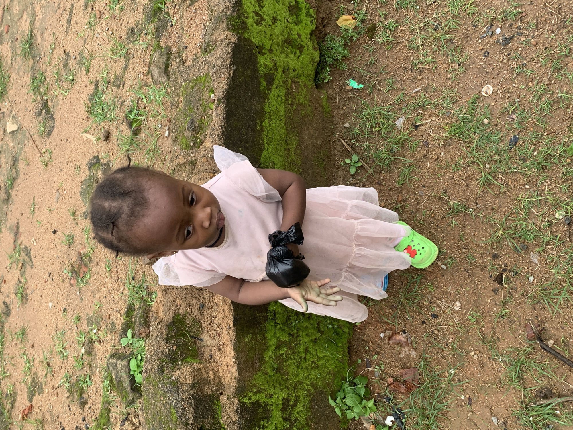 An undated, eye-level, full shot shows a young African child standing on dirt in Barakin Lerawi, Nigeria, holding a black plastic bag. The child, with dark skin and tightly pulled-back hair, wears a light pink, ruffled dress and bright green crocs. The child's left hand holds the black plastic bag, and their right hand is partially visible. The background features a low, moss-covered concrete wall and a dirt ground with sparse green grass and scattered debris. The overall lighting suggests an overcast day.