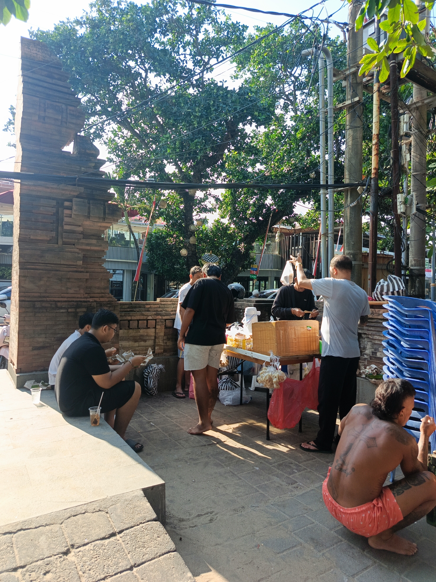 In Kuta, Indonesia, a group of people are gathered outdoors, possibly near a street food vendor or a communal gathering area. The scene is set in daylight with a bright sun casting shadows. In the foreground, on the right, a person with extensive back tattoos is squatting, facing away from the camera, wearing red patterned shorts. To the left, two men are seated on a concrete ledge. One man, wearing glasses and a black t-shirt, is eating from a paper container. Another man in a white t-shirt is seated next to him.
In the mid-ground, a table is set up with food items, including what appears to be packaged food and possibly some snacks. Several people are interacting around this table. One man in a black t-shirt and shorts is standing, appearing to be involved in the transaction or distribution of food. Another man in a light blue t-shirt is reaching towards the table with his right arm extended, holding a plastic bag. Behind them, a stack of blue plastic chairs is visible, suggesting a public space or an event.
The environment includes a large, mature tree with lush green foliage providing shade. There is also a decorative brick structure on the left, resembling a traditional Balinese archway or entrance. Electrical wires and poles are prominent in the background, indicating an urban setting. The ground is paved with interlocking bricks. The overall atmosphere suggests a casual, communal gathering in a public space on a sunny day.