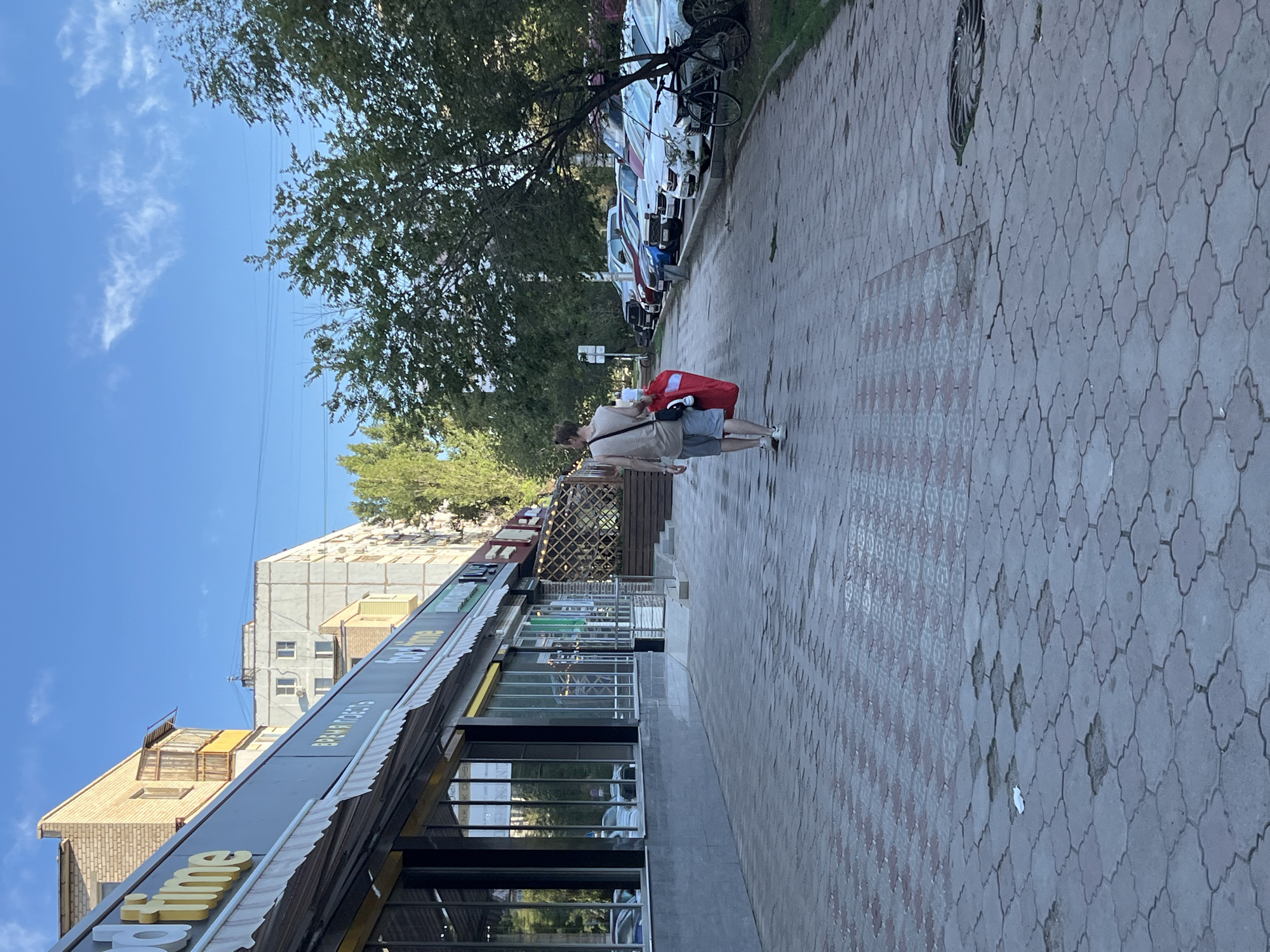 A man walks away from the camera on a sidewalk in Astrakhan, Russia, carrying a large red bag and wearing a light-colored tank top and grey shorts. The sidewalk is paved with interlocking grey and red stone tiles, some of which are damaged. To the left of the sidewalk is a row of shops with awnings and large glass windows. The shops have signs that read "food time" and "Время ПоЕсть" (Vremya PoEst' - Time to Eat in Russian). To the right of the sidewalk is a row of parked cars and trees. The sky is blue with a few wispy clouds.
