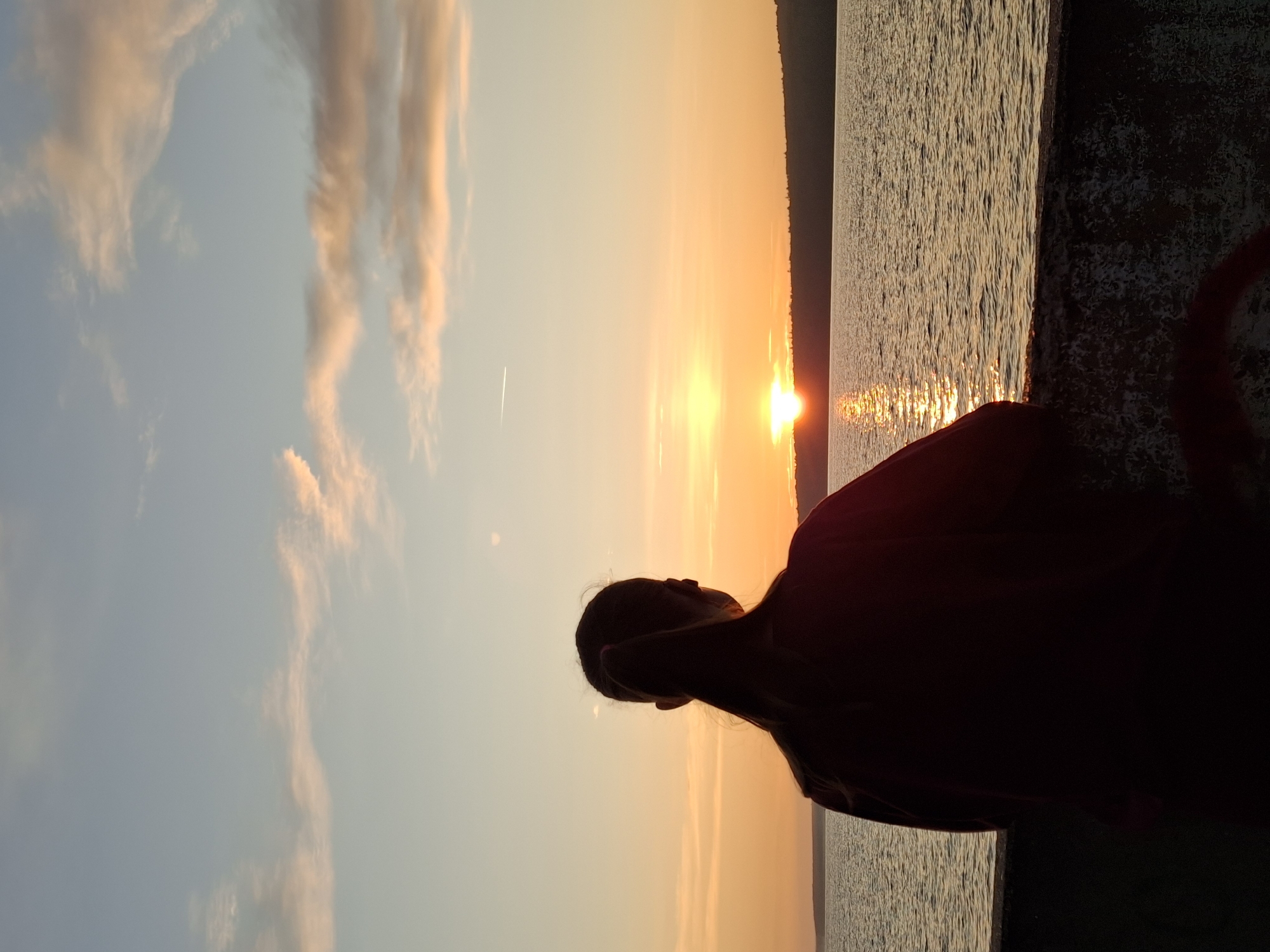 In the city of Chaikovsky, Russia, a person with their back to the camera stands on a stone barrier, looking out at a serene body of water as the sun sets on the horizon. The sky is painted with warm hues of orange and yellow, with scattered clouds tinged with pink. The sun, a bright orb, is just above the distant tree line, casting a shimmering path of light across the water. The person is silhouetted against the vibrant sky, their hair tied back in a ponytail. The overall scene evokes a sense of peace and contemplation.
