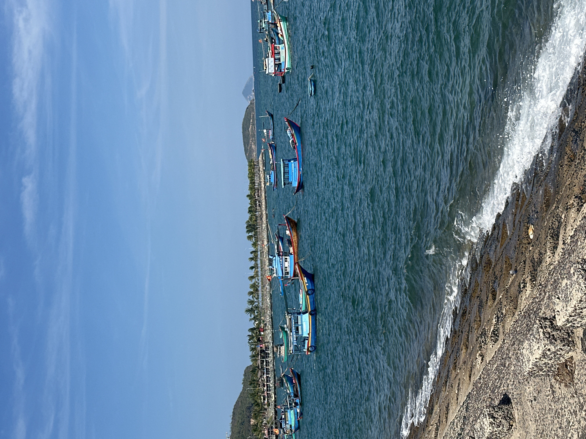 The image depicts a coastal scene in Bac Nha Trang Ward, Vietnam. Several colorful fishing boats are moored along the coastline. The water is a deep blue, with white waves crashing against a rocky embankment. A line of palm trees and low-lying hills run along the shore. The sky is clear and blue with thin white clouds.