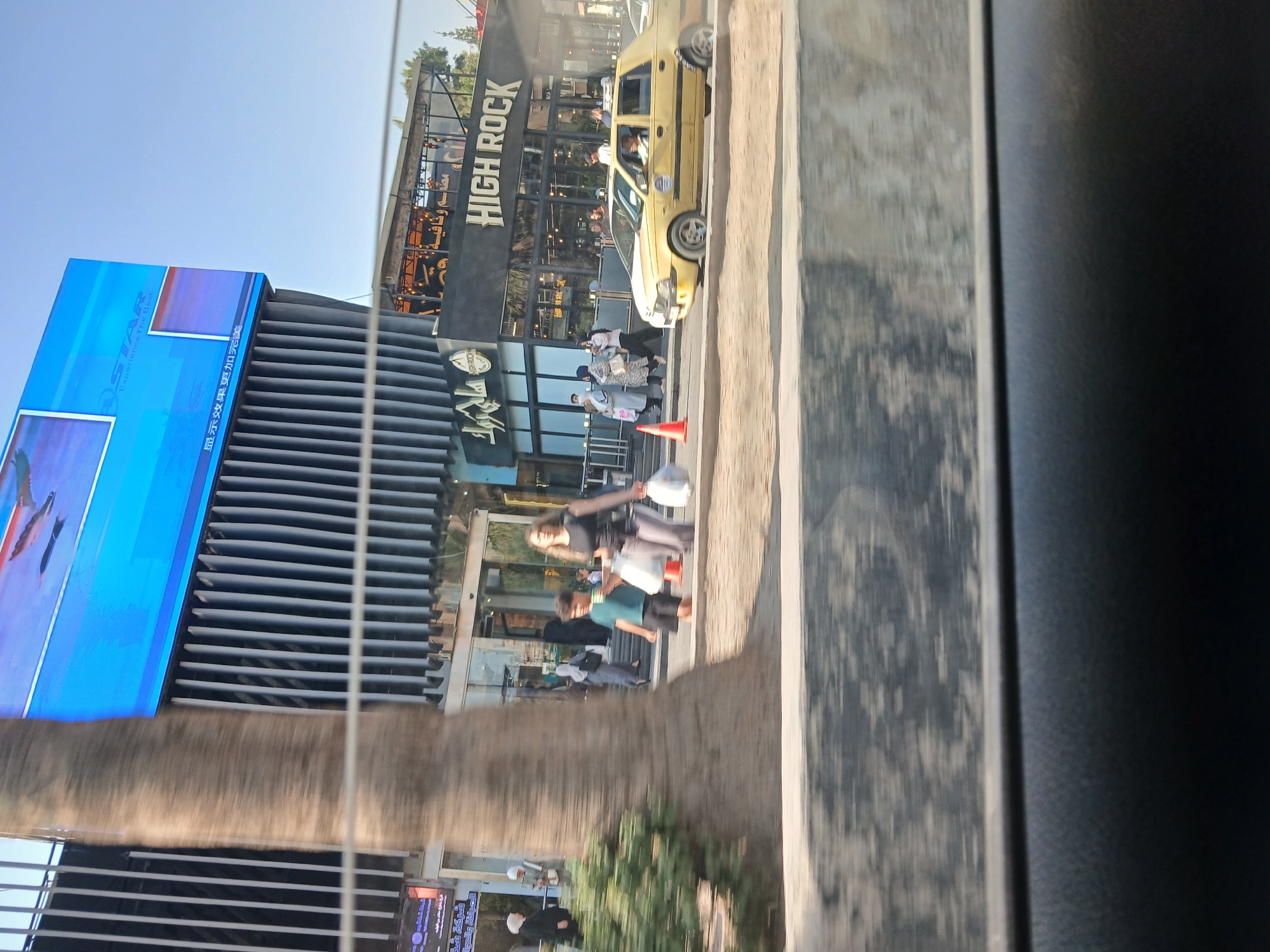 The image, taken from a vehicle, depicts a street scene in Barza Municipality, Syria. In the foreground, a large tree trunk is visible on the left. Behind it, a group of pedestrians are walking. A young boy in a teal t-shirt and dark shorts walks towards the right, carrying white bags. To his right, a woman with dark hair, wearing a black top and gray pants, also carries white bags. Further back, several other people are seen walking or standing. A yellow taxi is driving on the road.
The background features a modern building with a large blue digital screen displaying advertisements. The building has a ribbed facade. To the right of this building, there is a restaurant with a black facade and "HIGH ROCK" written in prominent white lettering. The restaurant appears to have an outdoor seating area with lights. The sky is clear and blue. The overall environment suggests a commercial area within the city.