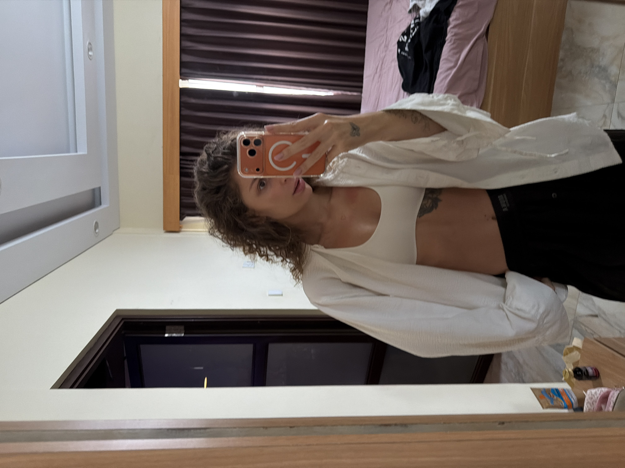 A young woman with long, curly brown hair is captured in a mirror selfie, holding an iPhone with an orange MagSafe case that partially obscures her face. She wears a white bralette or crop top, a loose white button-up shirt unbuttoned over it, and black bottoms. Visible tattoos include a butterfly on her right forearm/wrist and other designs on her left torso/ribcage. A watch is visible on her left wrist.
The setting appears to be an indoor room, likely a bedroom or dressing area, in North Kuta, Indonesia. Dark brown, horizontally textured curtains are drawn in the background, behind which a bed with light pink bedding and some black and white clothing items are visible. The clothing items on the bed appear to have white text, though it is illegible. The walls are light-colored (beige or off-white), and the ceiling features recessed lighting fixtures.
Reflected in the lower part of the mirror, a dark wooden door frame or entrance is visible to the left. To the right, on a surface beside the mirror, are several small objects: a small bottle or jar with a dark cap, a brown paper bag, and some folded textile items. The overall lighting suggests it is daytime, though the curtains are drawn. The image conveys a casual, posed moment.