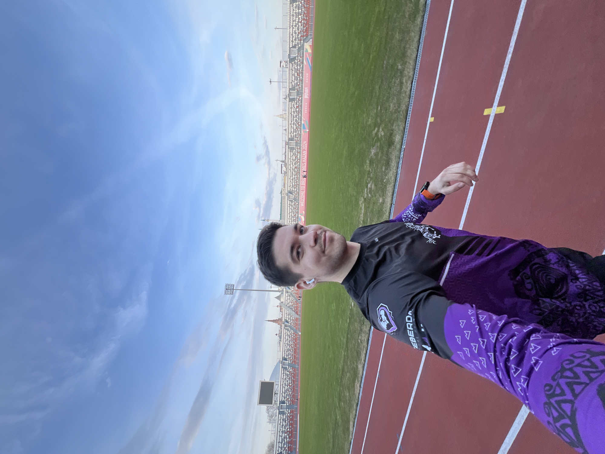 The image is a low-angle self-portrait of a man standing on a running track. He is wearing a black and purple athletic shirt with a logo on the left sleeve and matching purple leggings. He has an earbud in his left ear and a watch on his left wrist.
In the background, a stadium with rows of seats is visible. A green field stretches between the track and the stadium seating. Banners adorn the stadium with the text "WELCOME TO BRICS GAMES KAZAN 2024." In the distance, architecture resembling spires or towers can be seen. The sky is a mix of blue and clouds. The location appears to be a sports complex in Kazan, Russia, specifically the site of the 2024 BRICS Games.