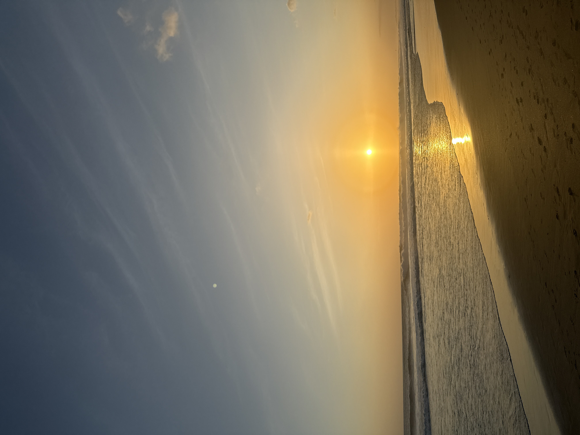 The image shows a serene sunset at a beach in North Kuta, Indonesia. The sky is a gradient of soft blues and oranges, with wispy clouds scattered across the horizon. The sun, a brilliant orb of golden light, is setting over the ocean, casting a shimmering reflection on the water. Gentle waves roll onto the sandy shore, leaving a wet sheen on the beach. Footprints are visible in the sand, suggesting recent human presence. The overall atmosphere is tranquil and peaceful.
