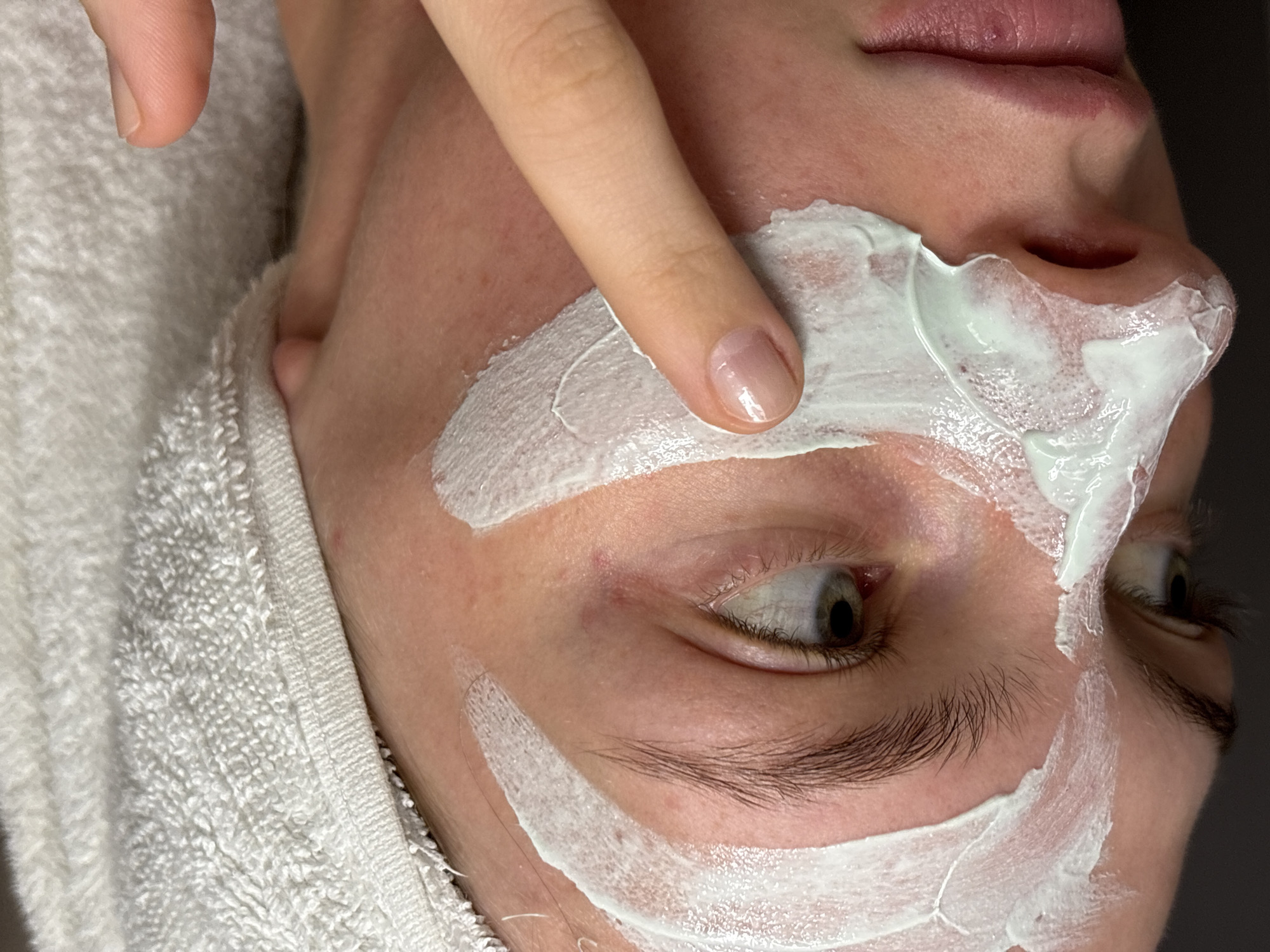 A close-up, high-angle shot captures a person receiving a facial treatment. The individual, likely female, is seen lying down with their head resting on a white terrycloth towel, which is visible on the left side of the frame. A hand, with natural, polished nails, gently applies or spreads a creamy, light green face mask onto the person's skin. The mask is visible in various patches across the forehead, around the eyes, cheeks, and extending towards the chin and nose area.
The person's face is largely in profile, with their eyes looking to the side, and a calm, neutral expression. Their lips are slightly parted, revealing a natural pink hue. Visible details include eyebrows, eyelashes, and natural skin texture beneath the mask. The lighting is soft but clear, highlighting the details of the skin and the texture of the mask. The background is a dark, indistinct blur, suggesting an indoor setting such as a spa, salon, or home. There is no visible text within the image. The scene depicts a moment of skincare or beauty routine.