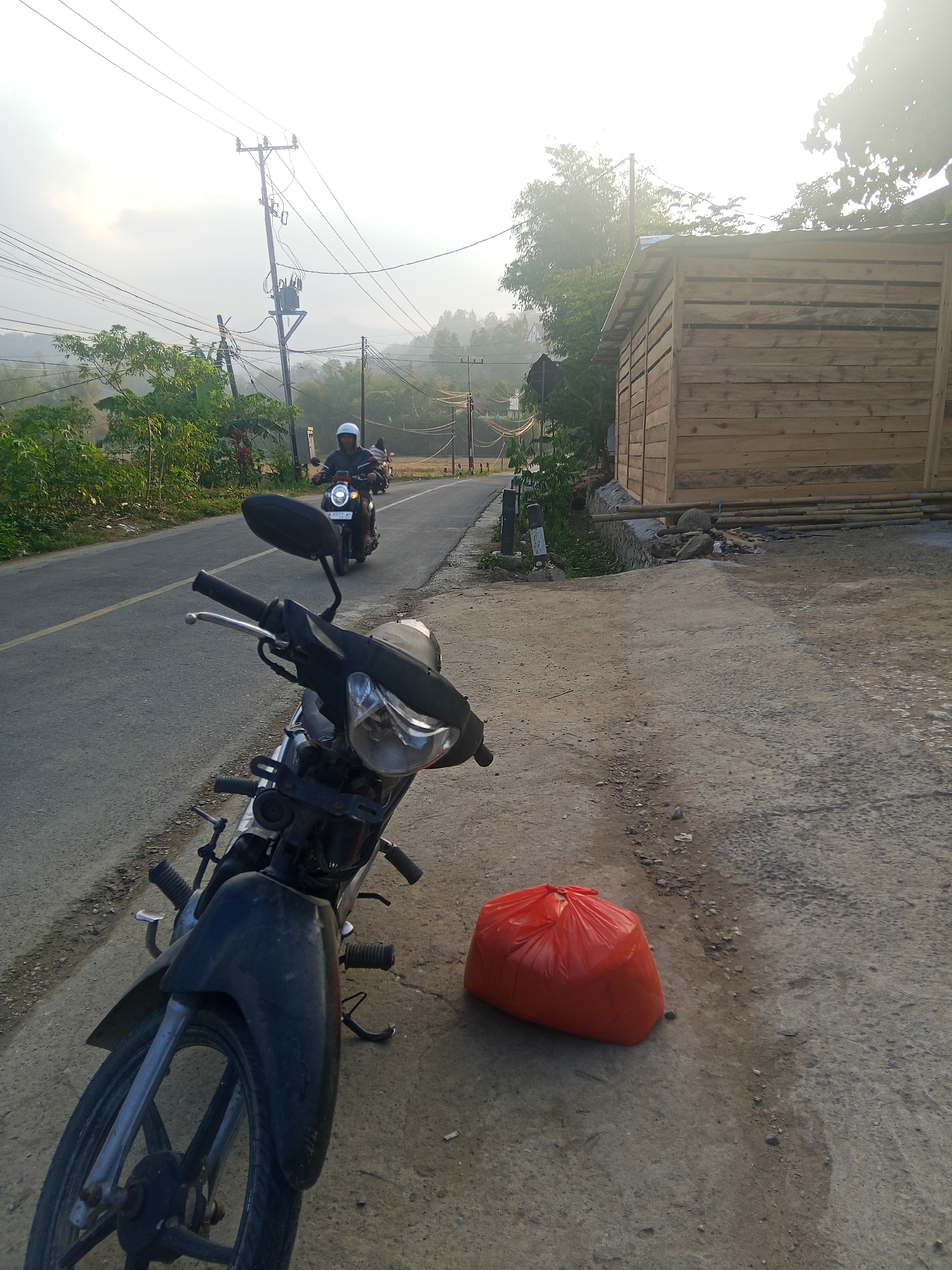 This image depicts a roadside scene in Batu Papan, Indonesia, under a hazy, overcast sky. In the foreground, a black motorcycle is parked on a rough, gray asphalt shoulder. A bright orange plastic bag lies on the ground near the motorcycle. The road curves to the left in the background, with two people on motorcycles riding away from the camera. One person is wearing a white helmet. To the right of the road, a newly constructed wooden building with a metal roof stands. Lush green trees and vegetation line the road and the background hills. Utility poles with numerous wires are also visible along the roadside. The overall lighting suggests it is either early morning or late afternoon.