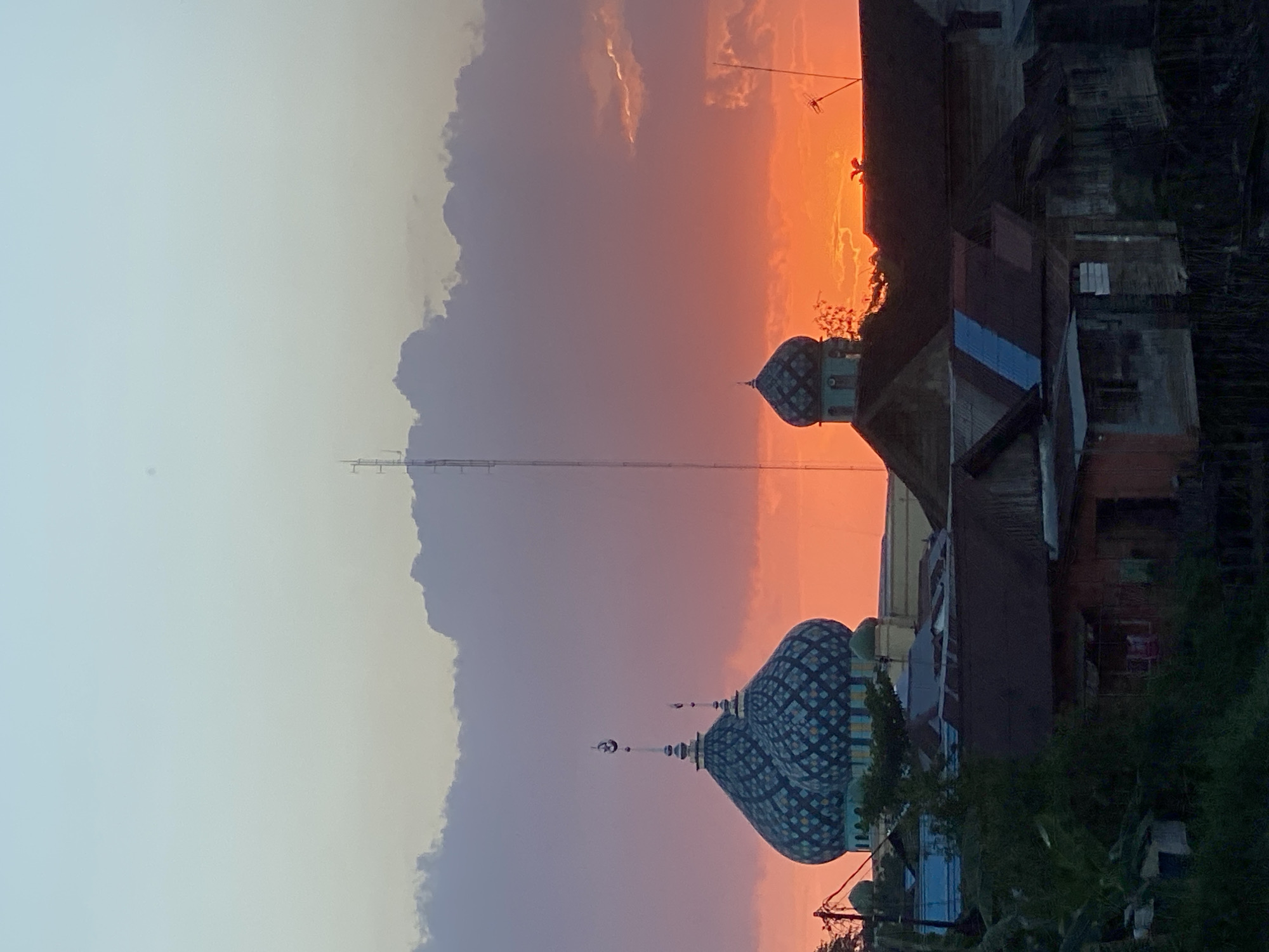 The image shows a sunset over the city of Tunggul Irang Ulu, Indonesia. The sky is a vibrant orange and pink, with clouds scattered throughout. In the foreground, there are several buildings, including two mosques with blue and green domes. There are also trees and other vegetation in the foreground. The scene is peaceful and serene, with a sense of tranquility.