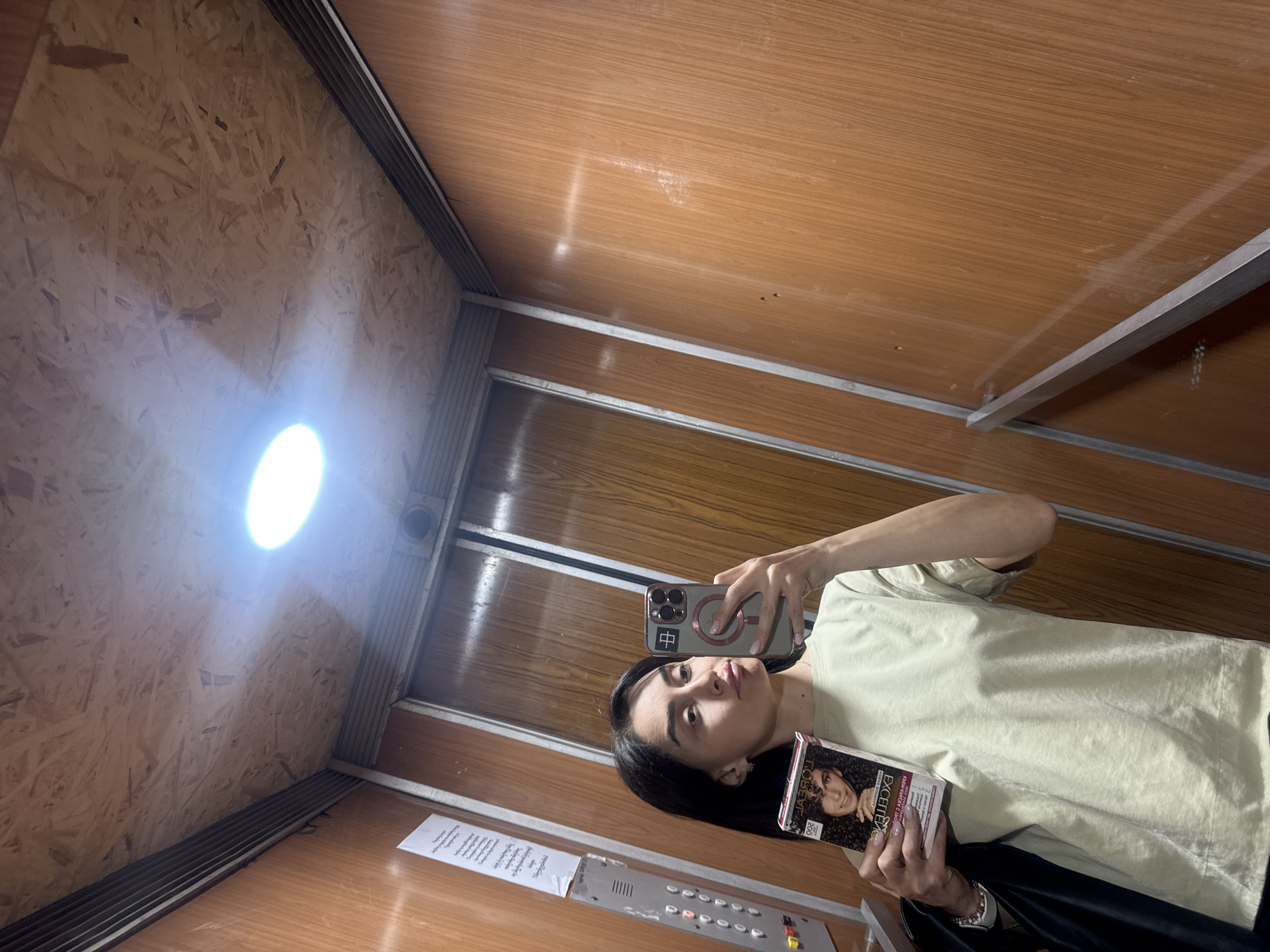 An indoor, eye-level selfie shows a person reflected in an elevator, which serves as the setting. The elevator interior features predominantly light brown wooden paneling on the walls and ceiling, with some sections possibly made of particle board or OSB on the left wall (from the selfie's perspective). Silver metallic strips accent the panel edges and joints. A bright, round artificial light source is prominent on the ceiling.
The subject is a young individual with dark hair, wearing a light-colored (possibly pale green or beige) long-sleeved top and dark bottoms. They are holding a grey/silver smartphone, encased in a clear magnetic case with a prominent red/orange Chinese character "中" (zhōng) on the back, to take the reflection selfie. In their left hand, they hold a rectangular product box for "L'ORÉAL EXCELLENCE" hair color, displaying a model with dark, curly hair and the number "200," along with some smaller Cyrillic text.
The person has a neutral to slightly pouty expression, looking towards the camera. Below them, an elevator control panel with various colored round buttons is visible. A white piece of paper with unreadable, likely Georgian or Russian, text is affixed to the wall near the control panel. The overall impression is that of a casual selfie taken in a functional, albeit somewhat utilitarian, elevator in Tbilisi, Georgia, during what appears to be daytime indoors.