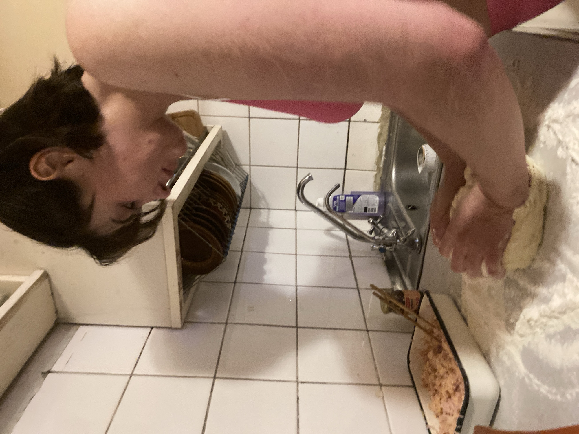 A person is kneading dough on a flour-dusted countertop in a kitchen. The kitchen has white tiled walls and a stainless steel sink with a faucet and a bottle of dish soap. A small dish of pink filling with chopsticks is to the left of the person. The person is wearing a pink top.