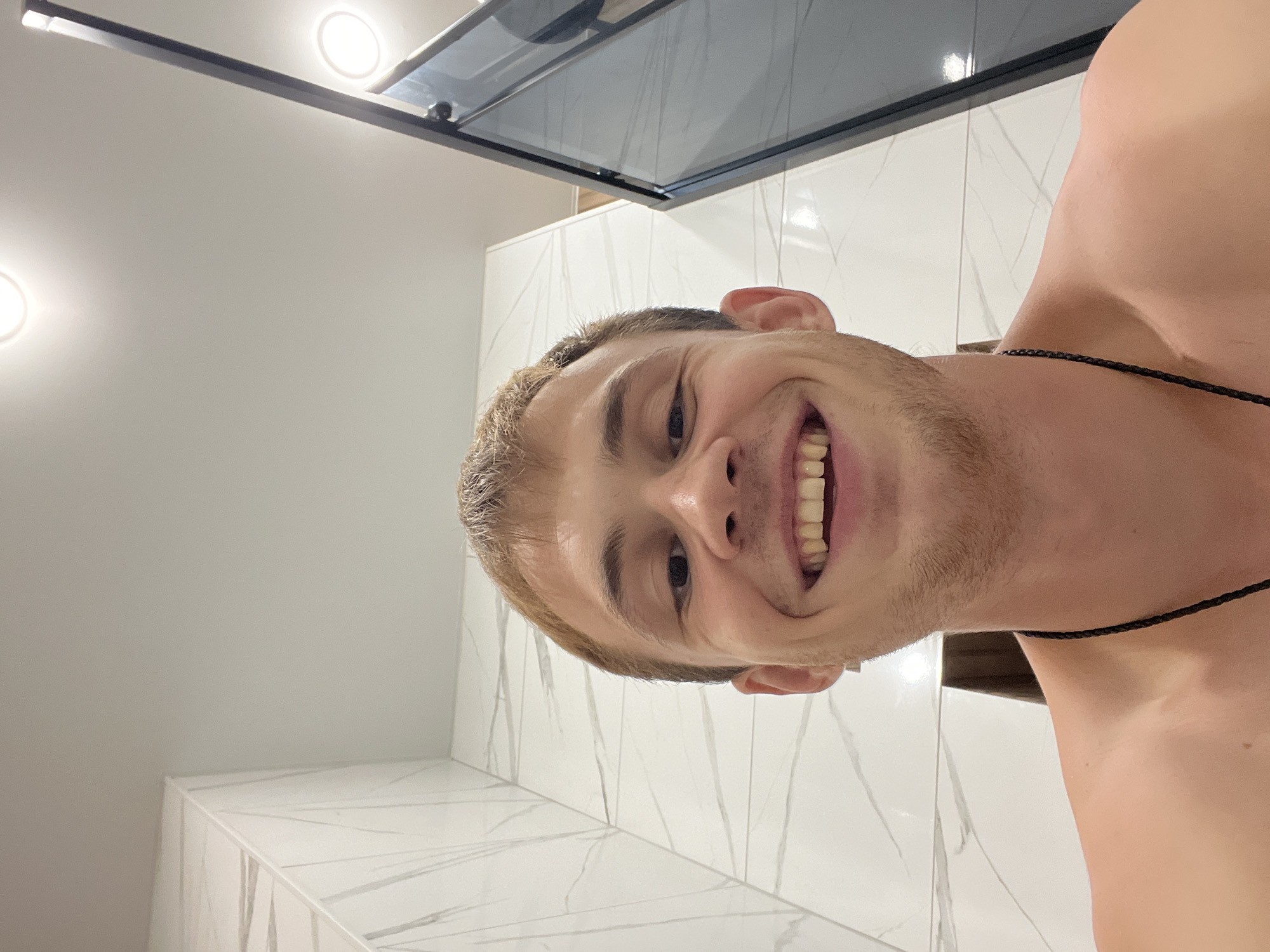 A young man with short, light brown hair and a wide smile is pictured in a close-up selfie. He is shirtless, and a black cord necklace is visible around his neck. He is in a bathroom with white marble tiles on the walls and a glass shower door. Two circular lights are visible on the ceiling. The overall lighting is bright and airy.