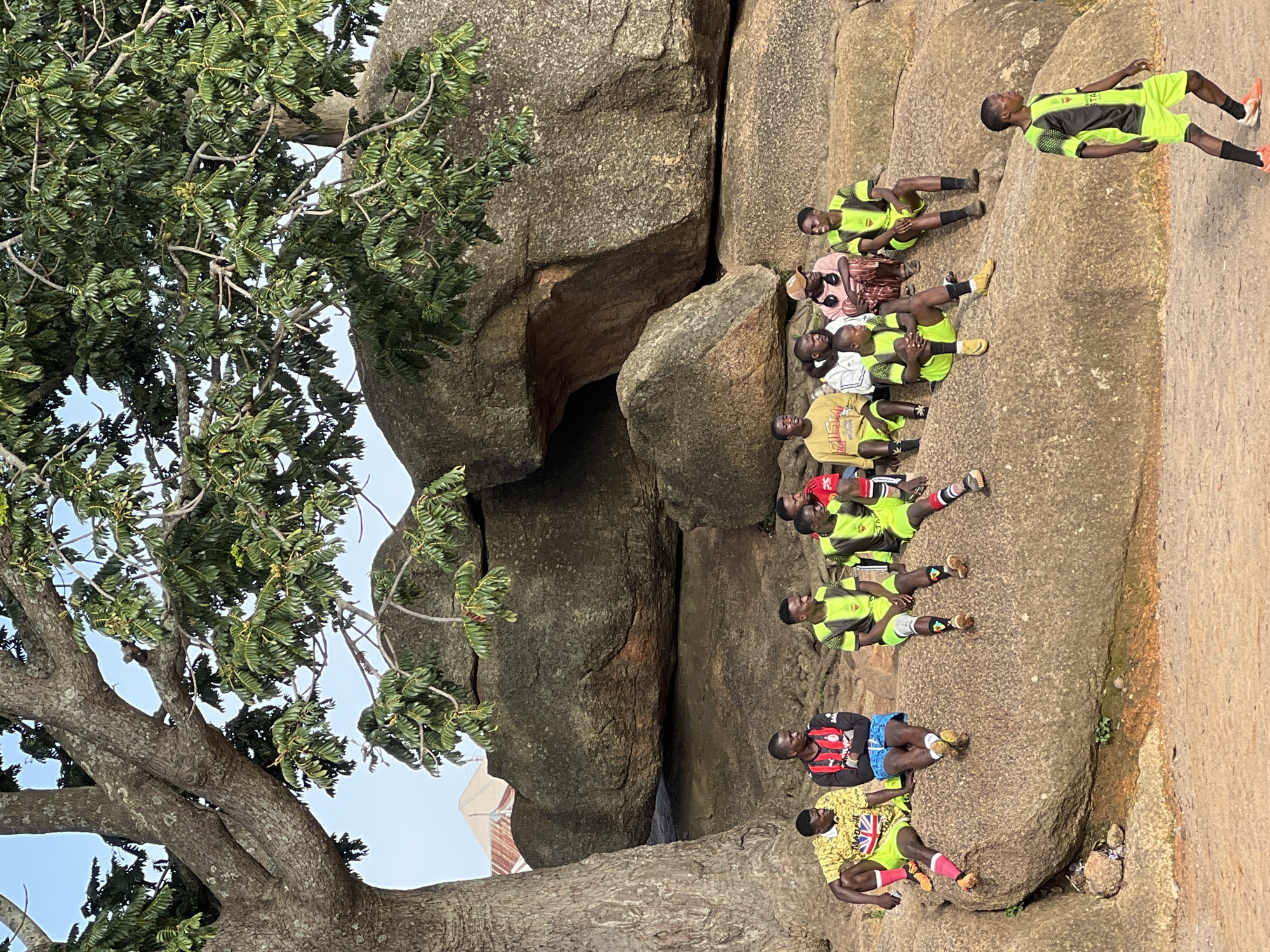 In the city of Jos, Nigeria, a group of young men, most of whom appear to be dressed in soccer attire, are gathered on a large rock formation under the shade of a massive tree. The formation of boulders creates a natural amphitheater-like setting. Several individuals are seated on the rocks, facing towards the left side of the frame, engaged in conversation. One young man in a bright yellow and white patterned shirt and lime green shorts, with the Union Jack flag design visible on his shorts, sits prominently on the left. Beside him, a man in a red and black soccer jersey and blue shorts is seated. Further to the right, several other men are wearing lime green and black soccer jerseys and shorts. One person is wearing a light pink garment and a cap, appearing to be an adult, possibly overseeing the group. Another young man in a similar lime green and black soccer uniform stands to the right, facing away from the seated group and towards the foreground, his stance suggesting he is about to engage in an activity or has just finished. The environment is rocky and arid, with sparse vegetation growing around the rocks and at the base of the large tree. The background reveals more of the rugged, rocky terrain, with hints of man-made structures, possibly buildings with corrugated roofs, in the distance under a clear sky. The overall scene depicts a casual gathering of young men in an outdoor, natural setting.