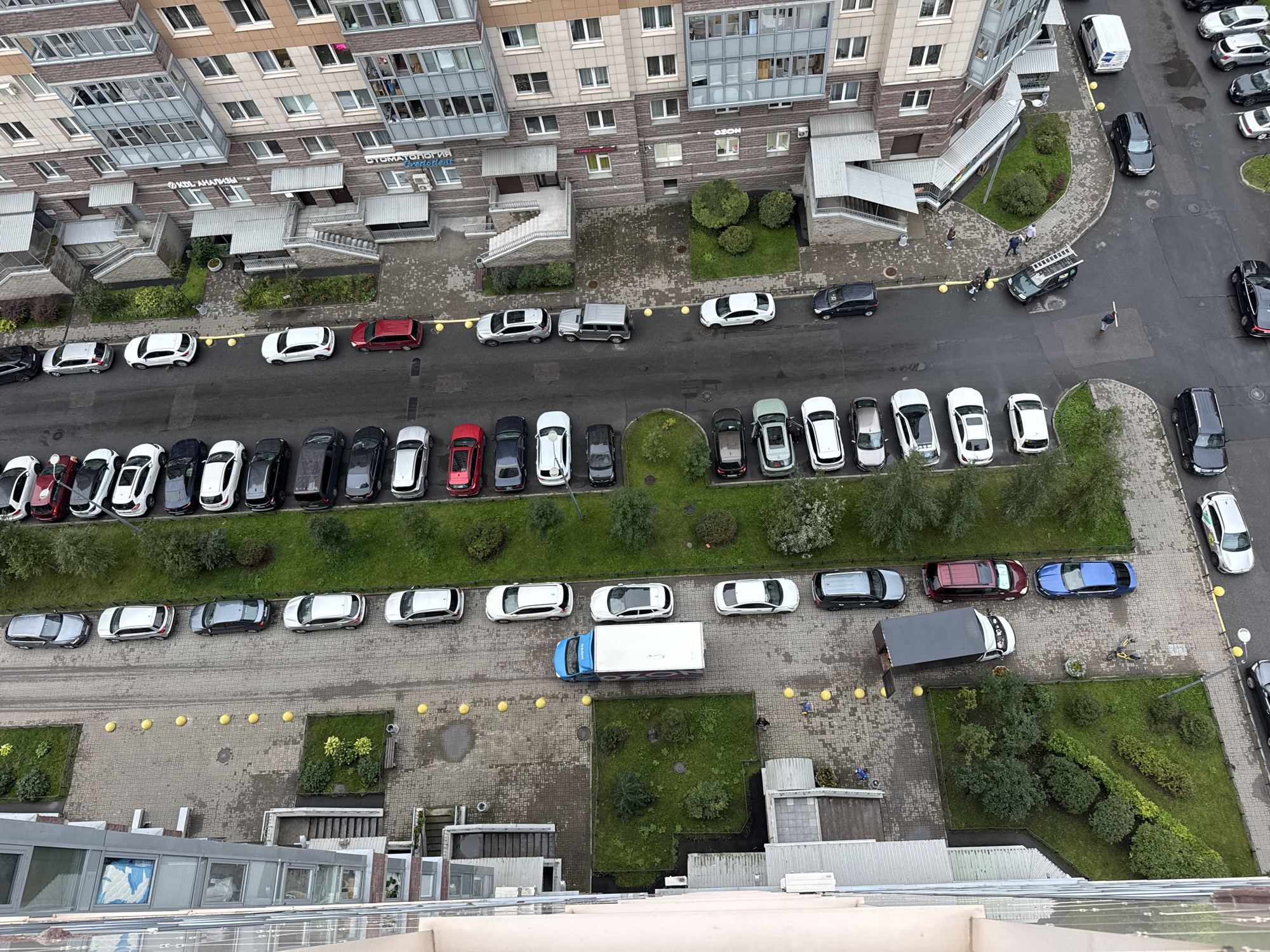 This is an aerial, eye-level, full-shot photograph of a residential courtyard in Sinyavino, Russia. The image captures a row of parked cars, apartment buildings, and green spaces. In the foreground, a multi-story apartment building with glass balconies dominates the left side of the frame. Opposite the building, a paved courtyard area features yellow bollards and several parked cars, including a blue truck. Further back, more apartment buildings are visible, along with patches of grass and trees. The ground is a mix of paved areas and grass. The sky is overcast. Several individuals are scattered throughout the courtyard, some walking and others standing near the parked vehicles.