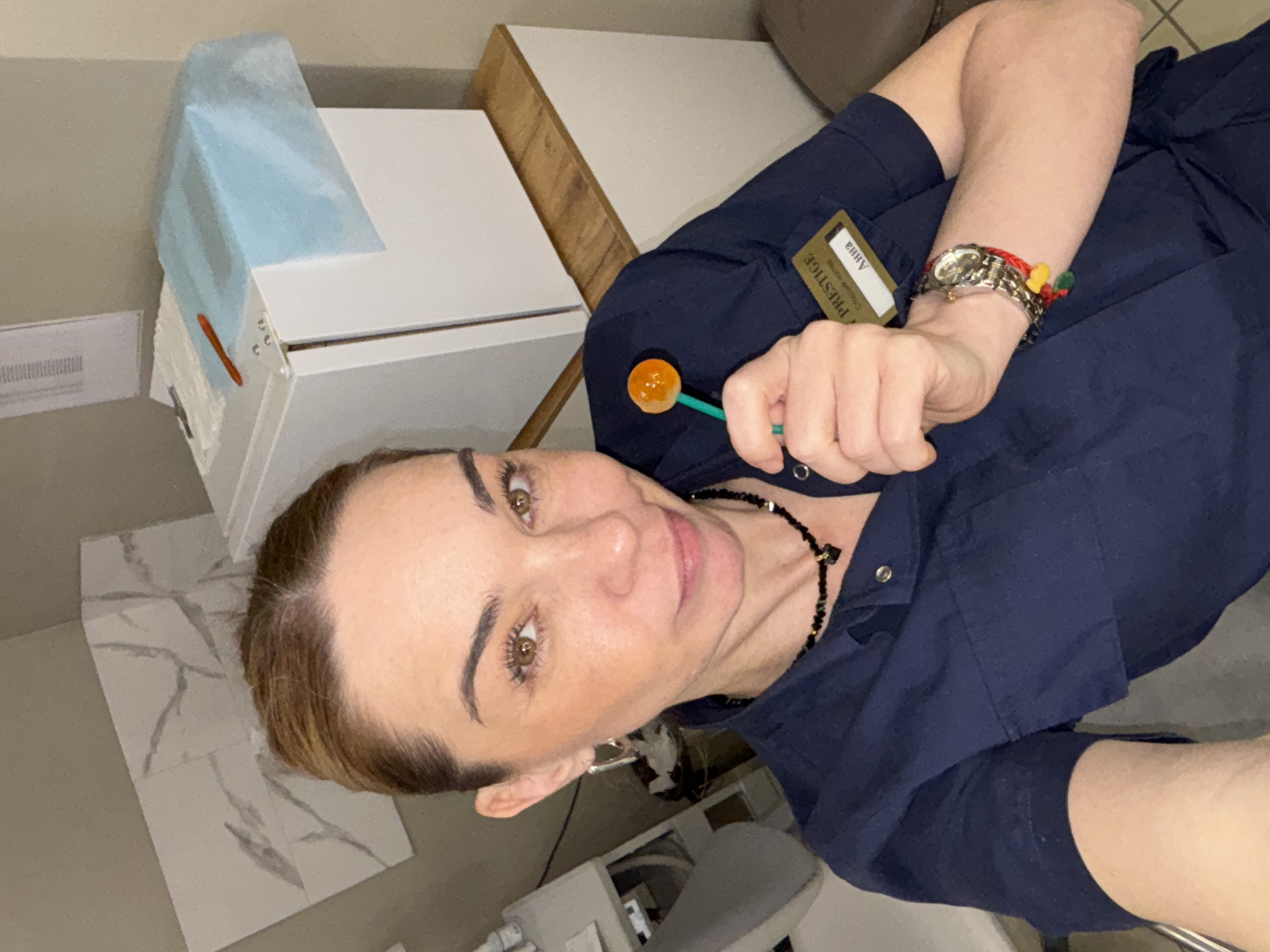 A woman in a dark blue uniform stands in what appears to be a medical or beauty treatment room, based on the equipment visible in the background. She holds a lollipop in her right hand and wears a silver wristwatch and a bracelet with small colorful pom-poms on her left wrist. Her uniform has a name tag that reads "Anna Prestige". She is wearing a black beaded necklace. The room has neutral-toned walls and marble-patterned floor tiles. A cabinet with a blue cloth or cover is visible in the background. The specific location within Vsevolozhsk, Russia cannot be determined from this image alone.