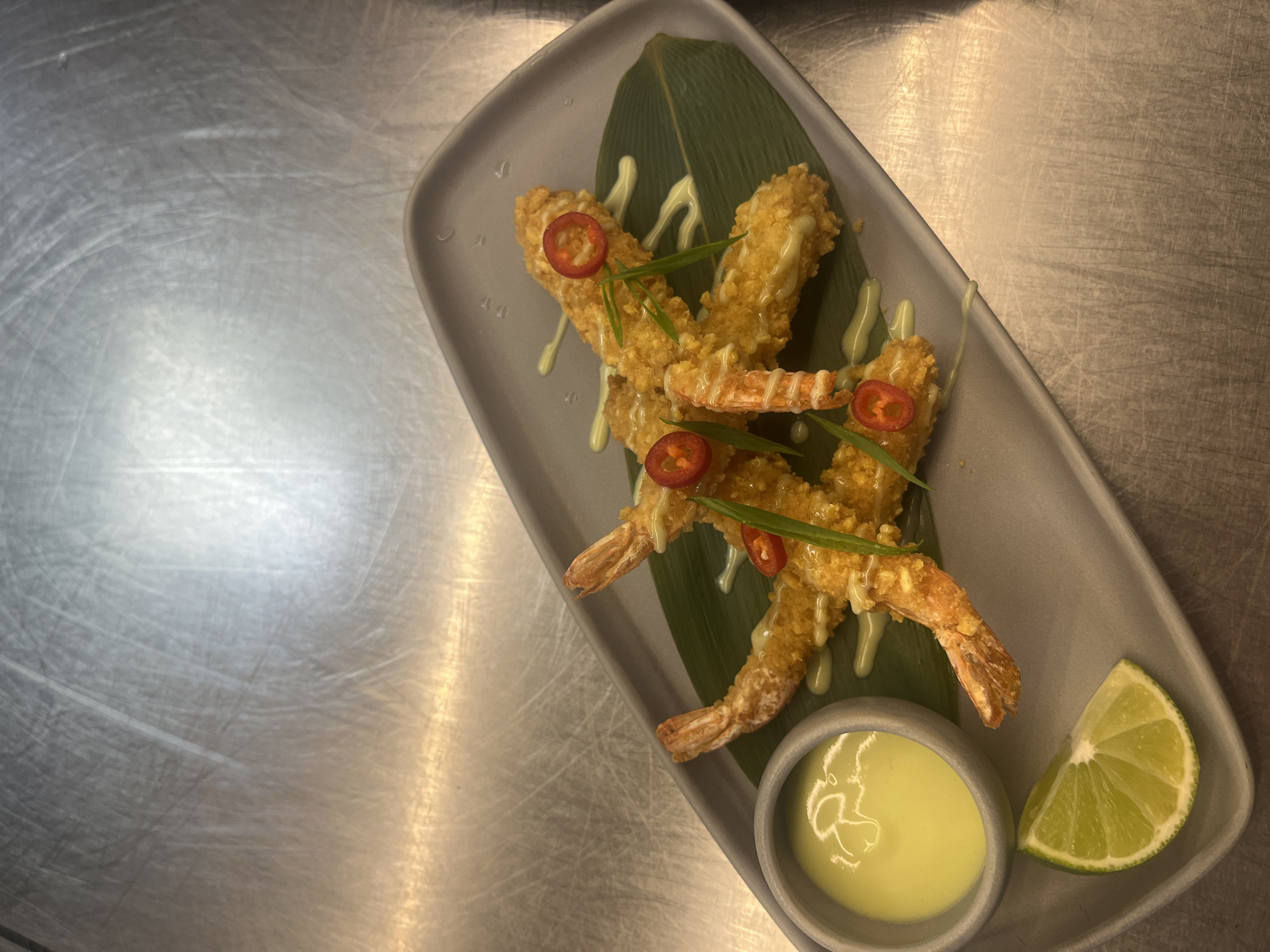 The image displays an overhead view of a prepared food dish, likely served in a restaurant setting. The main subject is a grey, irregularly shaped serving plate resting on a metallic, scratched surface, which resembles a stainless steel kitchen counter or serving area.
On the plate, several golden-brown, breaded shrimp are arranged artfully on a large, dark green leaf, possibly a banana leaf, serving as a decorative and textural base. The shrimp appear to be deep-fried, possibly Panko-crusted, and are generously drizzled with a creamy, light green-yellow sauce, suggesting an aioli or a spicy mayonnaise. Garnishing the shrimp are thin slices of bright red chili peppers and delicate green strips, likely chives or spring onions, which add vibrant color and freshness to the presentation.
Accompanying the shrimp on the plate is a small, round grey bowl filled with more of the light green-yellow dipping sauce. Adjacent to this, a fresh, vivid green lime wedge sits, offering a citrusy accent to the meal.
The metallic background surface is highly reflective, showing numerous scuffs and scratches, which reinforce the impression of a professional culinary environment. The lighting is bright and even, casting subtle reflections on the shiny surfaces. There are no people visible, nor any discernible activity beyond the static presentation of the dish. No visible text is present in the image. The meticulous plating suggests the dish is ready for consumption or photography, possibly from a restaurant in Sofia, Bulgaria.