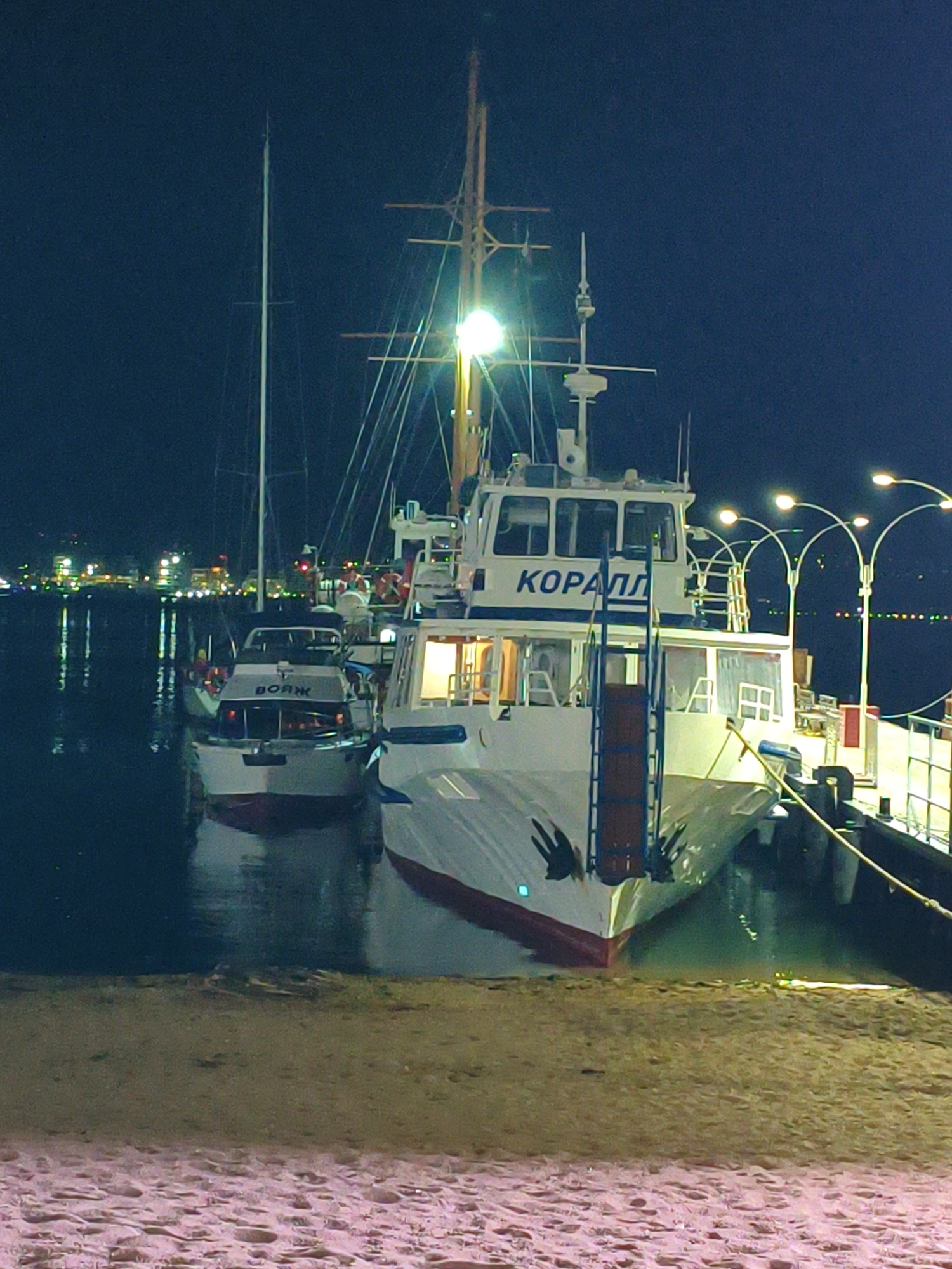 A nighttime view shows a harbor with two boats docked. The larger boat, in the foreground, is white with a red stripe along its hull and has the word "КОРАЛЛ" written in blue on its side. It has multiple decks and a mast. The smaller boat, to the left, is also white and has the word "ВОЯЖ" written on its stern. Both boats are illuminated by artificial lights from the shore and streetlights on the pier. The water is dark and reflects the lights. In the background, a city skyline with numerous lights is visible across the water. A sandy beach or shoreline is in the immediate foreground, with the sand appearing pinkish due to the lighting. The overall scene depicts a calm night at a harbor.