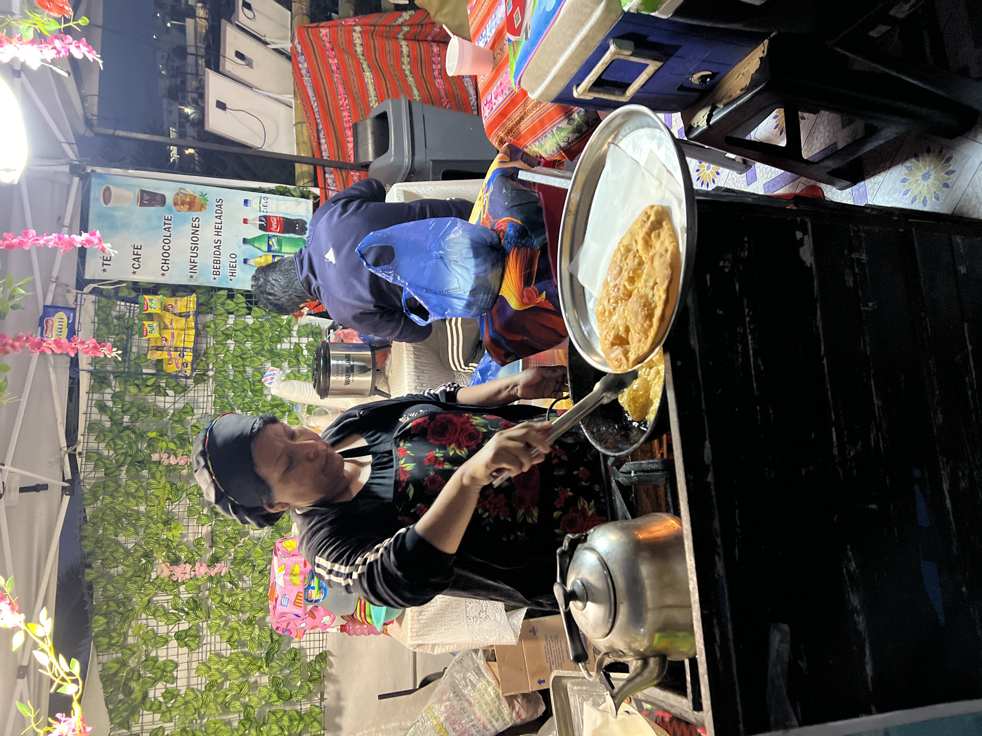 Here's a detailed description of the image:
The image captures a street food vendor in Arica, Chile, preparing and selling fried bread, possibly sopaipillas or a similar traditional snack.
A woman with dark hair pulled back and wearing a black headscarf is seen cooking. She has on a black jacket with white stripes on the sleeves and a floral-patterned apron. She is using tongs to transfer fried bread from a pan to a silver tray lined with paper towels.
Behind her, another person is seated. It's hard to see specific details about them, but they are wearing a blue jacket and have a blue plastic bag.
The food stand itself is constructed from dark-colored material, and a silver teapot sits in the foreground. The stall features decorations, including faux greenery and pink flowers. A sign lists the beverages available for sale, including tea, coffee, chocolate, infusions, and cold drinks, in Spanish. Bags of chips are also displayed for sale.