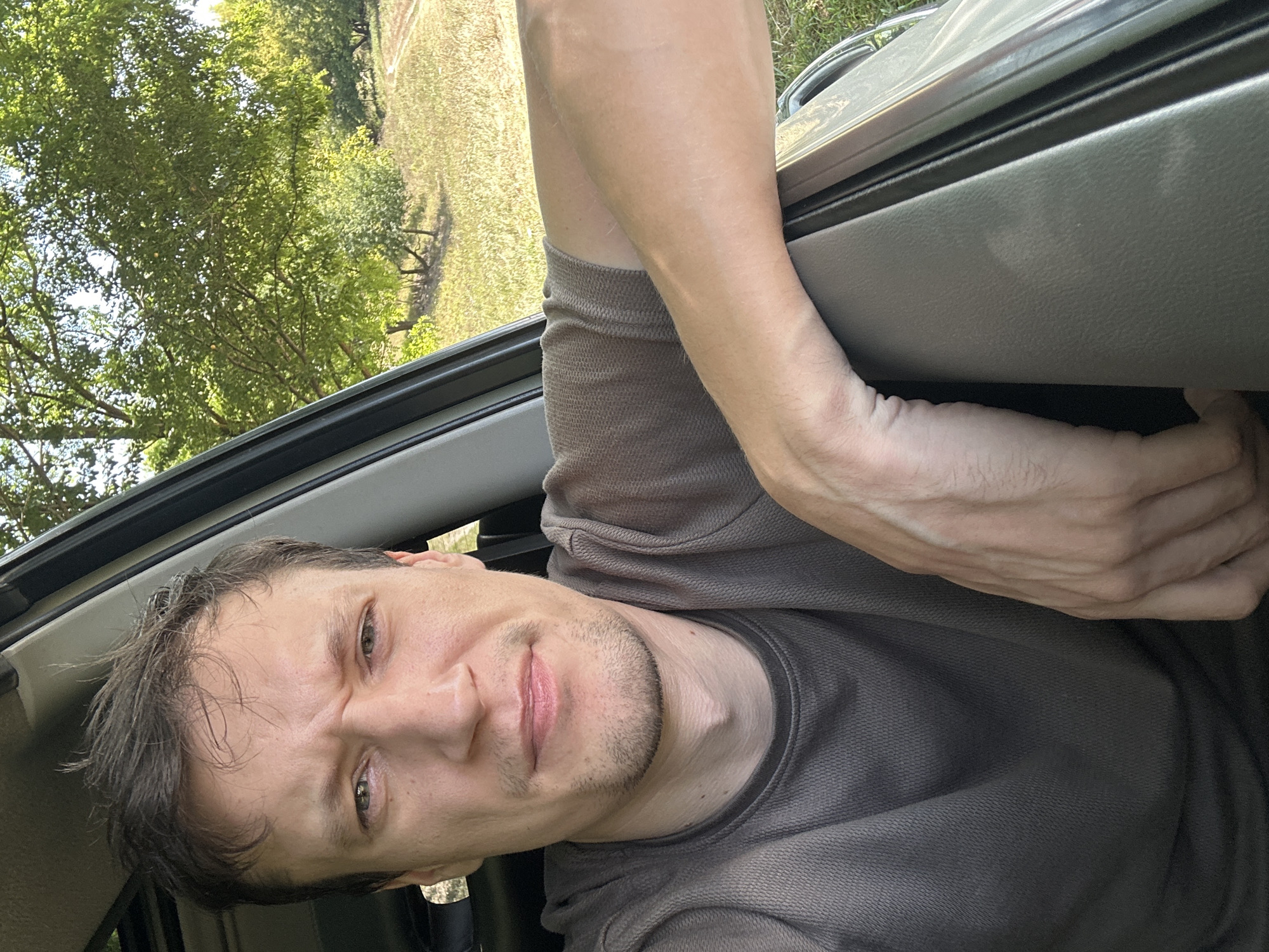 A man with short, dark brown hair and a light complexion is captured in a close-up, selfie-style shot from inside a vehicle. He is looking directly at the camera with a slightly furrowed brow and a neutral expression. He is wearing a dark grey, short-sleeved t-shirt. His left arm is resting on the car door, with his hand visible. The background outside the car window shows a grassy field with trees and a dirt road, indicating a rural or semi-rural setting, possibly on the outskirts of Makiv, Ukraine. The lighting suggests it is daytime, with sunlight filtering through the trees.
