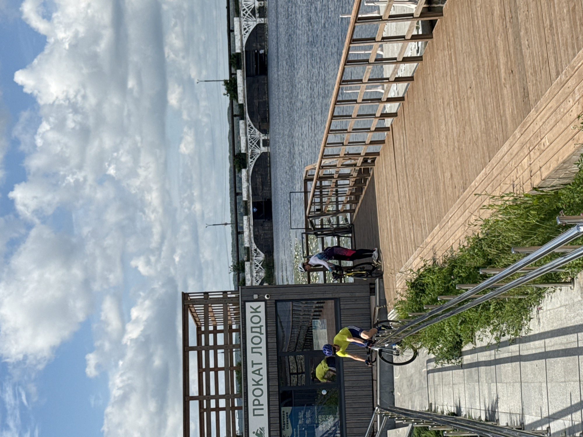The image depicts a scene at a boat rental facility, identified by the Russian text "ПРОКАТ ЛОДОК" (PROKAT LODOK) meaning "Boat Rental," located in Votkinsk, Russia. The facility has a wooden structure with a dark gray facade and a wooden railing. Two individuals are engaged in cycling on a wooden pier. One person, wearing a yellow shirt and blue helmet, is actively riding a bicycle down a tiled ramp with metal railings, moving towards the pier. Another person, in cycling attire with a white helmet, is standing next to a bicycle on the wooden pier, appearing to be pausing or preparing to ride. The pier extends over a body of water, likely the Votkinsk Reservoir, which is calm and reflects the cloudy blue sky. In the background, a large arched bridge, characteristic of infrastructure in Votkinsk, spans the water. The overall environment suggests a recreational area by the water, under a partially cloudy sky.