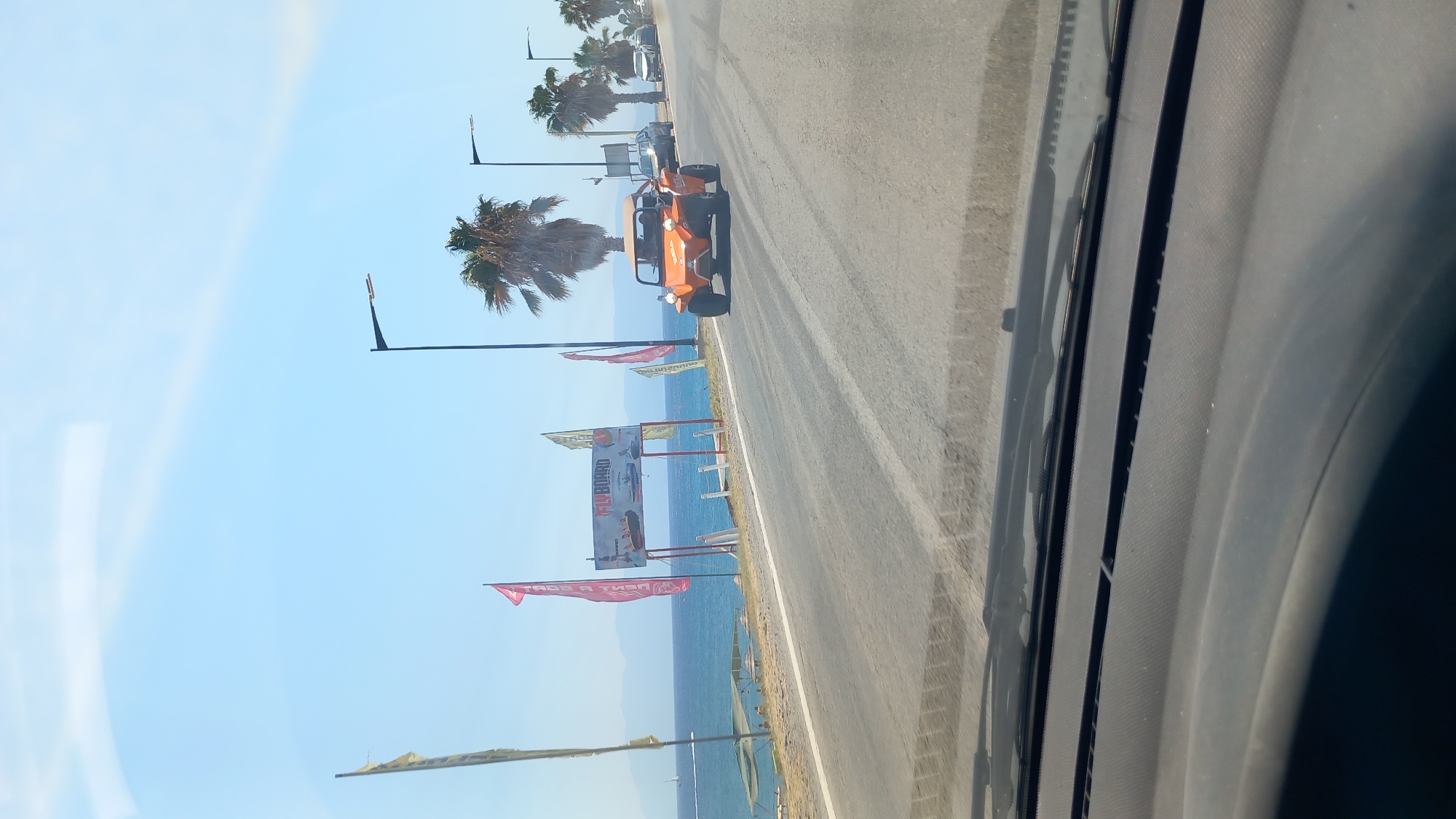 The image depicts a sunny day in Kos, Greece, likely along the coast. In the foreground, the dashboard and windshield of a vehicle are visible, suggesting the photo was taken from inside a car. The road curves to the right, with palm trees lining the side. On the left side of the road, an expanse of the Aegean Sea is visible, with mountains in the distance. Several flags are planted near the roadside, one of which displays "FLYBOARD". An orange dune buggy is driving on the road, with other cars visible further down the street. A few people are seen relaxing under umbrellas on the beach.