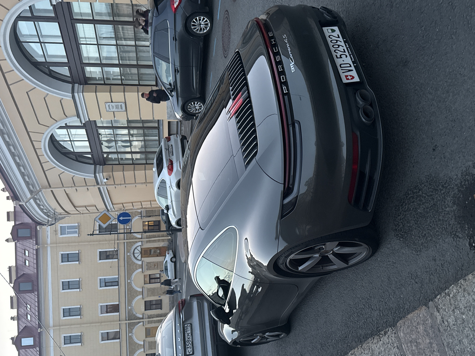 A dark grey Porsche 911 Carrera 4S is parked on a city street, angled towards the right. The car's rear is prominent in the foreground, displaying its license plate "VD-52992" and the text "PORSCHE ZENTRUM BASEL." Behind the Porsche, a dark grey hatchback and a silver sedan are also parked. In the background, a yellow building with arched windows and a mansard roof suggests an urban environment. A few pedestrians are visible walking on the sidewalk in front of the building. Traffic signs, including a directional arrow and a warning sign, are mounted on a pole near the building. The street appears to be wet, possibly from recent rain. The overall scene depicts a typical urban streetscape in Saint Petersburg, Russia.