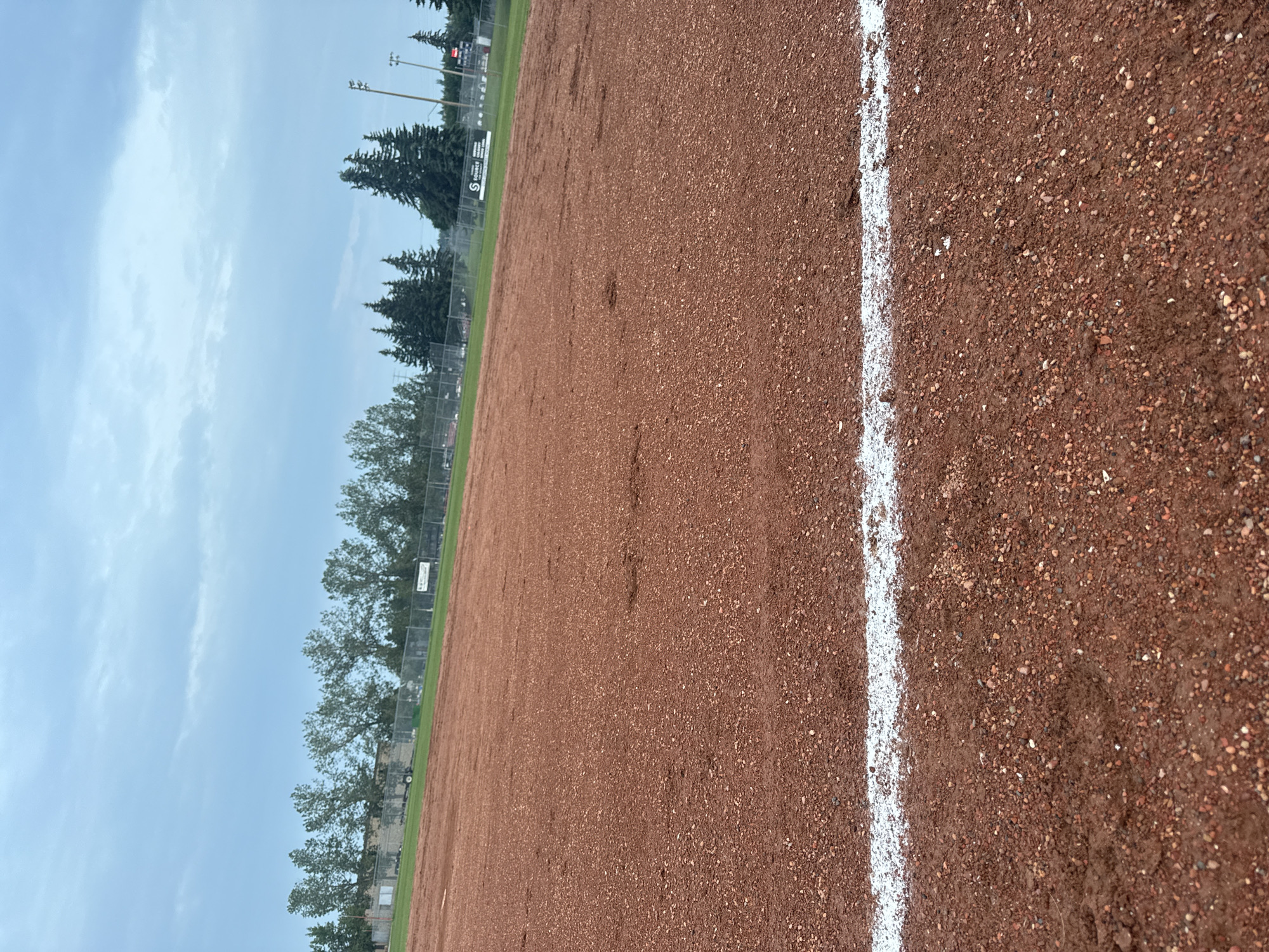 The image depicts an outdoor baseball field with a dirt infield and a grassy outfield, under a cloudy sky. A white line marks the edge of the infield in the foreground. In the background, a chain-link fence encloses the field, beyond which are trees, buildings, and sports lighting. The scene appears to be a public park or sports complex. No people are visible in the image.