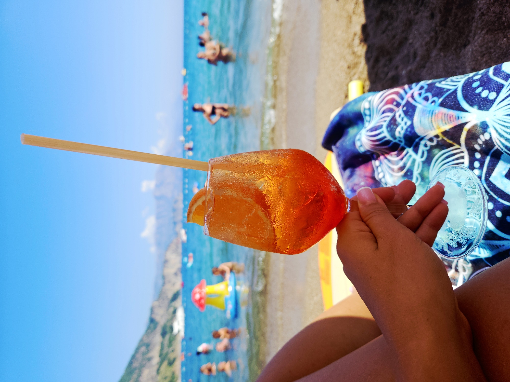 A woman's hand holds a glass of orange cocktail garnished with a slice of orange and a straw. The glass is beaded with condensation, indicating it is cold. In the background, the turquoise ocean stretches out to meet a hazy coastline and a clear blue sky. Several people are visible in the water, some swimming and others relaxing on inflatable toys. The beach in the foreground is dark sand, and a colorful patterned towel is draped over the woman's legs. The overall scene evokes a relaxed summer day at the beach.