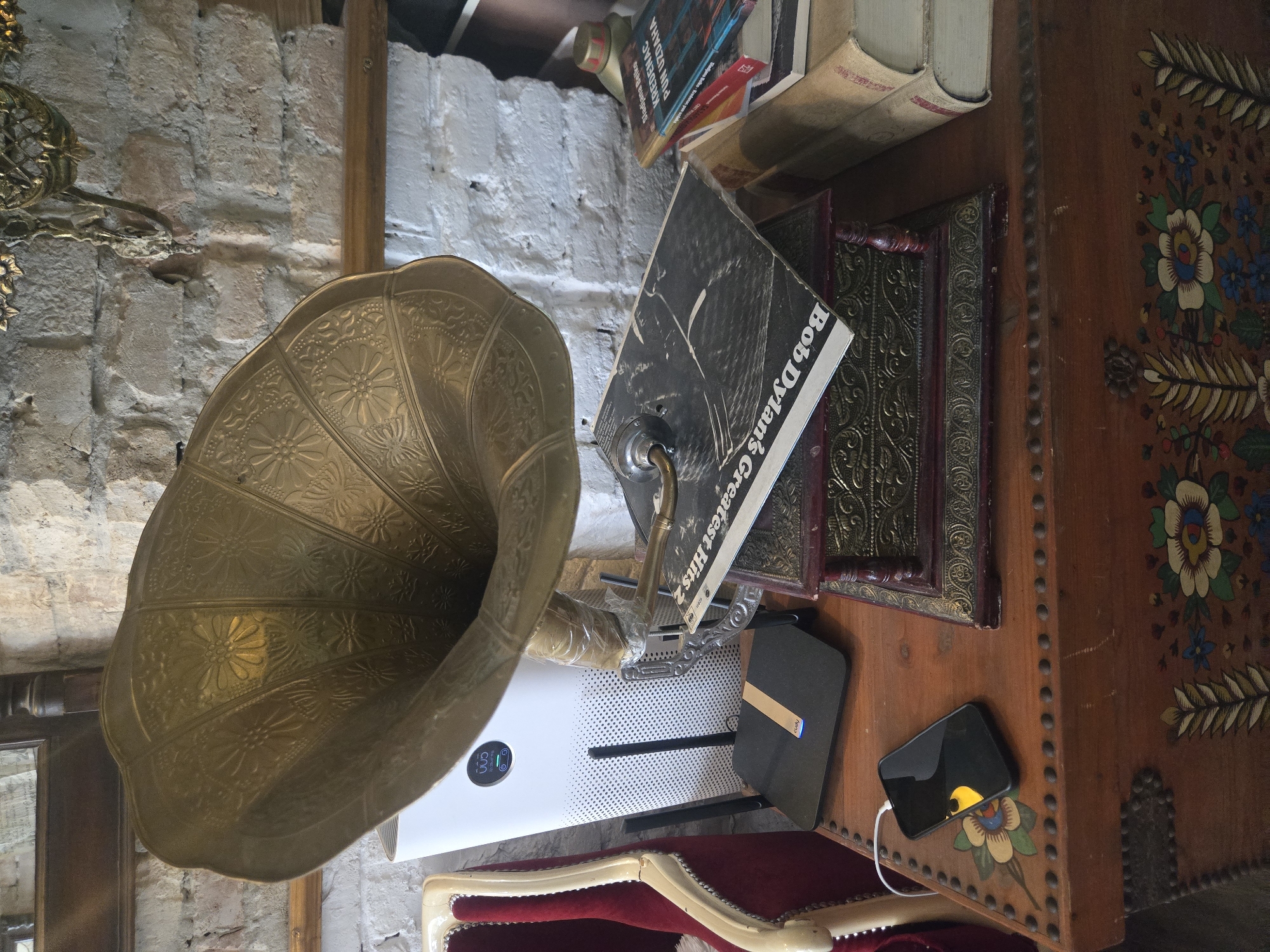 A still life scene depicts a vintage phonograph with a large brass horn, placed on a wooden chest or cabinet with floral folk art designs. A Bob Dylan record leans against the base of the phonograph. Next to it are several books and framed art. A modern air purifier with a digital display and a mobile phone charging nearby are also visible. The background shows a rustic brick wall, partially painted white. The scene's location is Palilula Urban Municipality, Serbia.