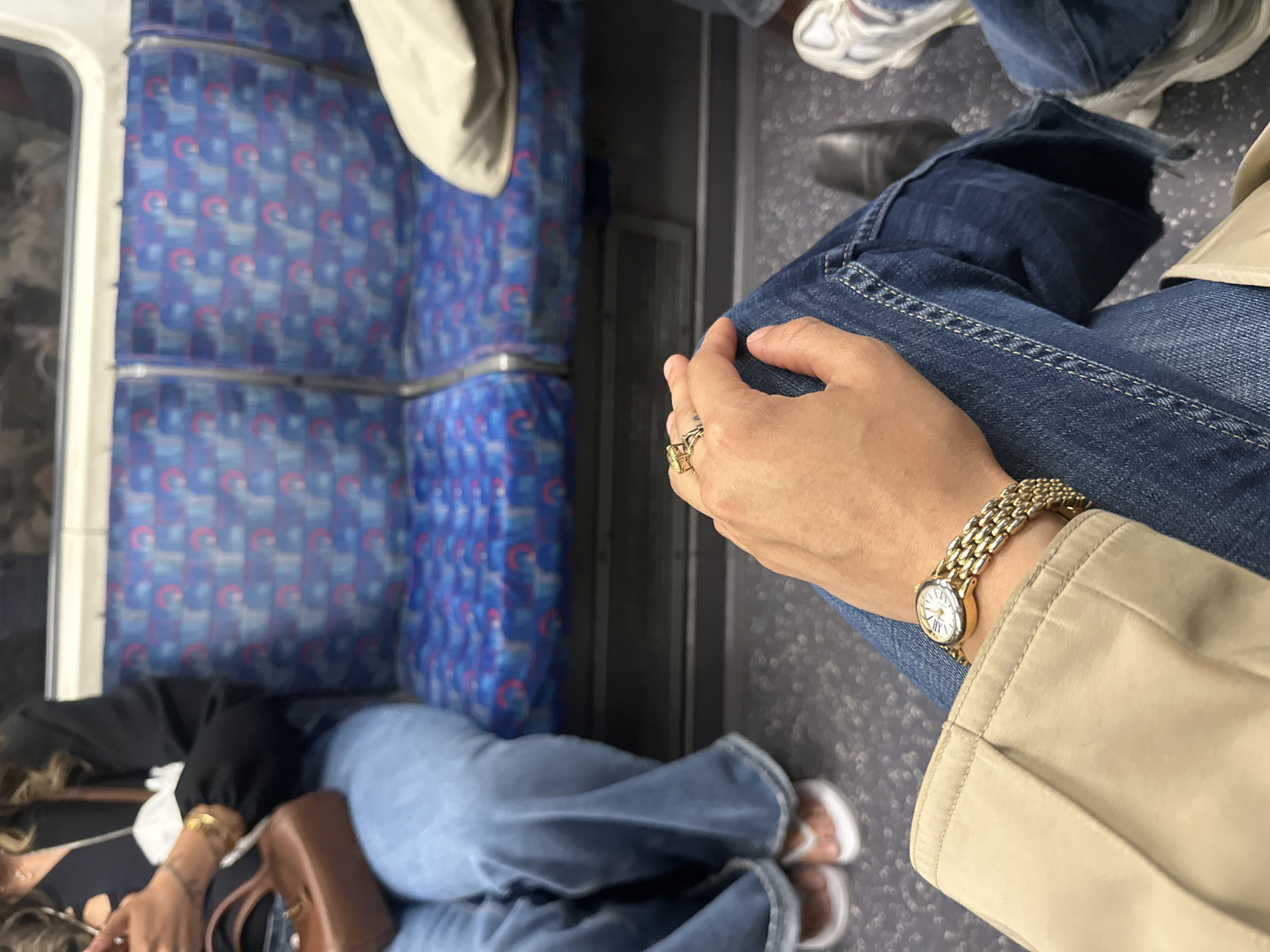 The image depicts an interior view within a London Underground train carriage in the United Kingdom. Distinctive blue patterned seat upholstery is visible. A person is seated, wearing jeans, a beige jacket, a gold watch, and a gold ring. Another individual appears to be lying down or asleep on the seat, with jeans, a black top, and a brown bag. There are at least two other passengers on board.