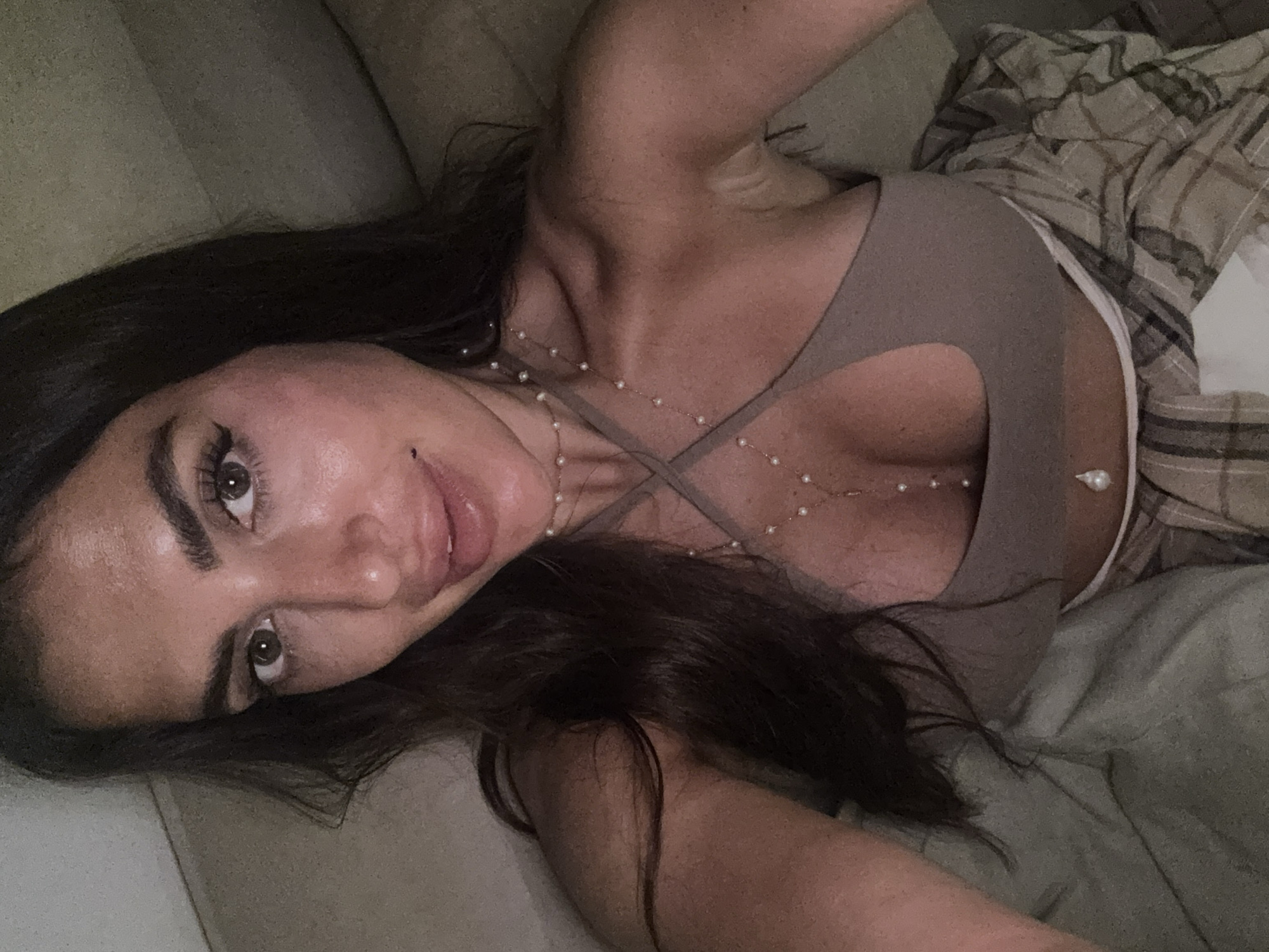 A woman is lying on a couch, facing the camera. She has dark hair, brown eyes, and light skin. She is wearing a light brown top with a cutout at the chest, and a pearl necklace. She is also wearing plaid pants. The background includes a tan couch and plaid sheets. The location is unknown; the presence of the city of North Kuta, Indonesia is not deducible from the image.
