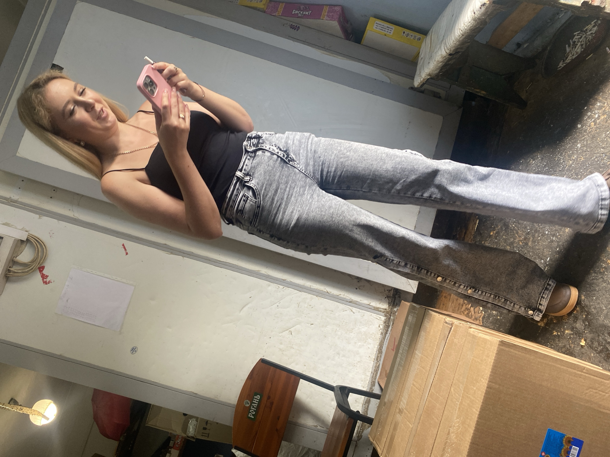 A young woman stands indoors, holding a pink smartphone and looking at it. She is wearing a black tank top and acid-wash jeans. In the background, a wooden chair with a logo that reads "РОГАНЬ" is visible. There are also cardboard boxes stacked nearby and a worn wooden bench. The environment appears to be some sort of storage area or workshop. The lighting suggests it is daytime, with a light source visible overhead.