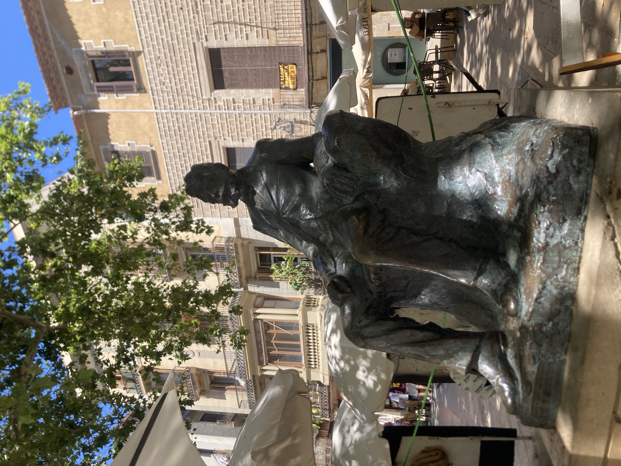 The photograph captures a bronze statue of a man with a beard and mustache, seated with his legs crossed. He is dressed in a suit, and his hands are resting on his lap. The statue is positioned in what appears to be a public square or plaza in Barcelona, Spain, as indicated by the architectural style of the surrounding buildings. The buildings are of a Mediterranean design, with ochre-colored facades, tiled roofs, and balconies with iron railings. Trees with green foliage are visible in the foreground and background, casting dappled shadows on the ground. White umbrellas from outdoor seating areas for cafes or restaurants are also prominent in the scene. In the background, several people are seen sitting at tables or walking, suggesting a lively atmosphere. A yellow sign with Spanish text is affixed to one of the buildings, possibly advertising a business. The sunlight indicates it is daytime, and the clear blue sky suggests fair weather.