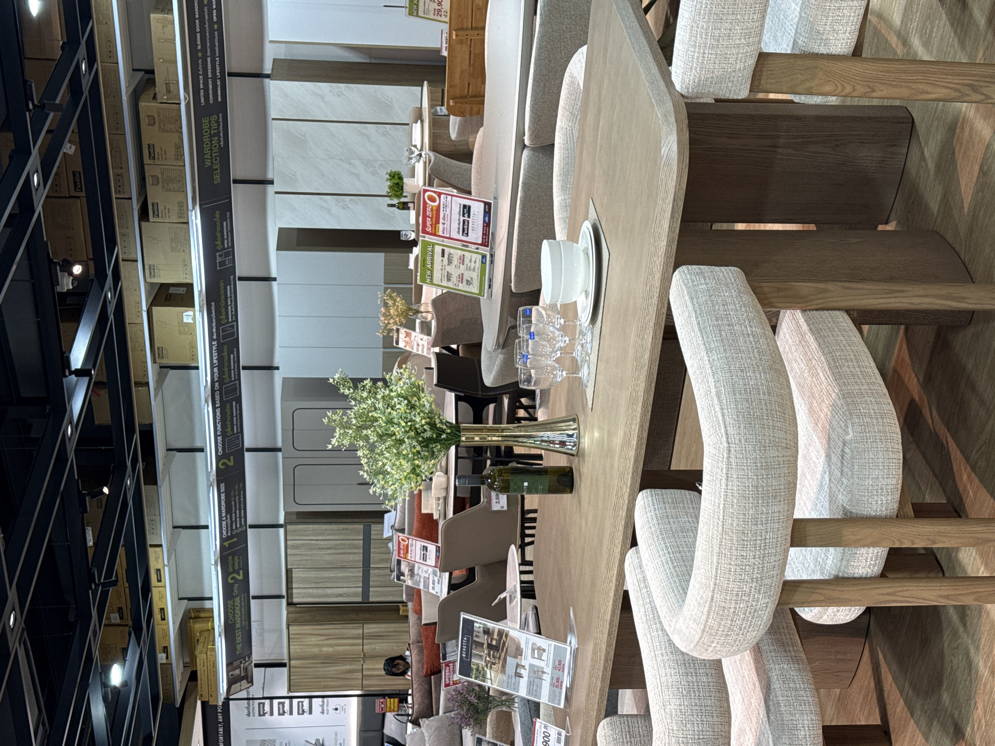 The image shows the interior of a furniture store. In the foreground, a light-colored wooden dining table is set with white plates, white bowls, and wine glasses. Two chairs with light-colored fabric upholstery and wooden legs are visible on either side of the table. A vase with green foliage is placed on the table, along with a green bottle of wine. In the background, more furniture, including sofas, chairs, and display shelves, are visible. Several signs are posted throughout the store. The store appears to be well-lit with overhead lighting. The overall scene suggests a retail environment where furniture is displayed for sale. Based on the context provided, this location is in Baan Chaweng Noi, Thailand.