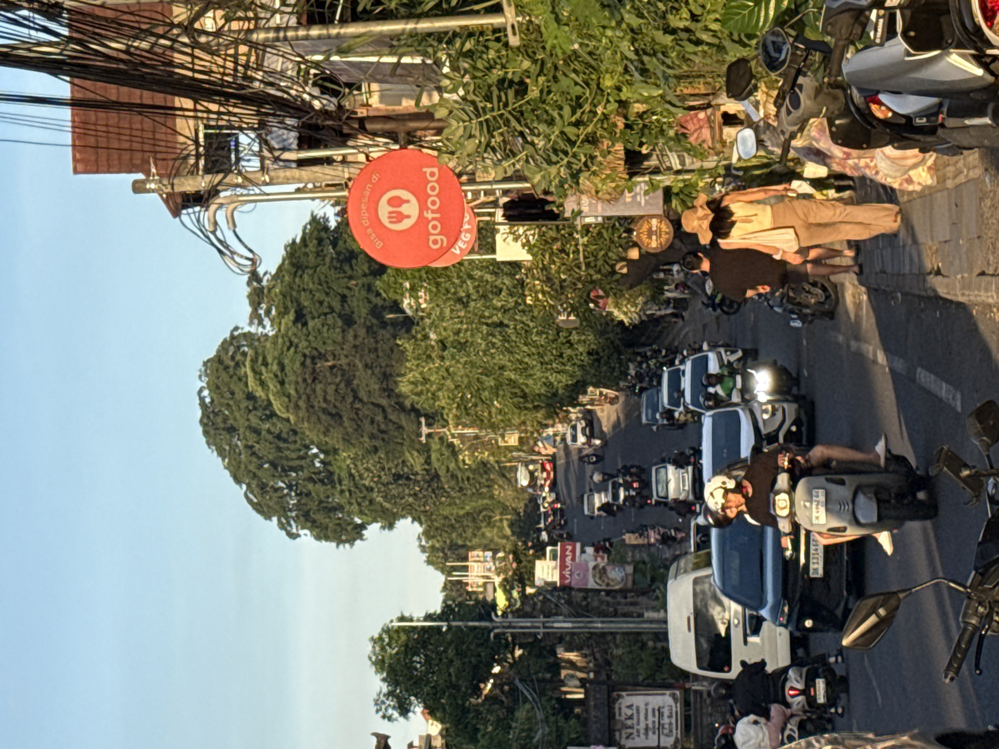 The image depicts a street scene in the Ubud District of Indonesia, characterized by heavy traffic including cars, motorcycles, and scooters. The road is lined with lush trees and vegetation. Prominently visible is a "GoFood" sign, indicating the presence of food delivery services. Overhead, there is a dense network of power lines. In the foreground, a couple is observed walking along the sidewalk adjacent to a parked motorcycle. The overall scene conveys a sense of everyday life in a bustling Southeast Asian urban setting.