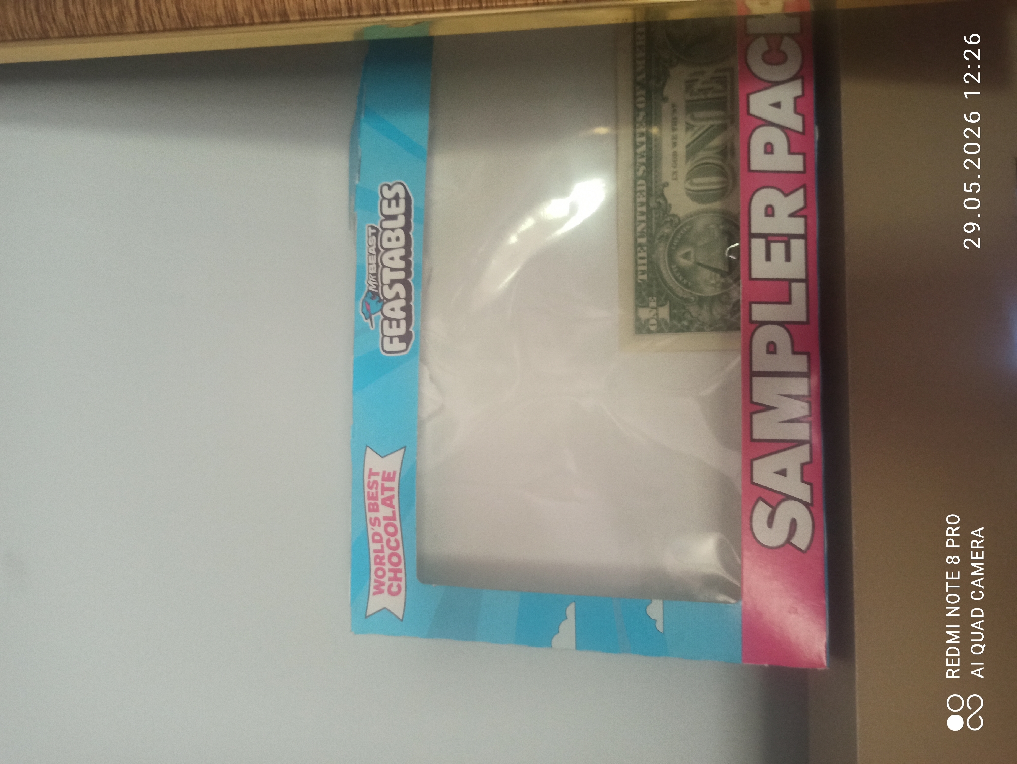 The image shows a MrBeast Feastables sampler pack box with a one-dollar bill partially visible through the packaging. The box is oriented vertically, with its front face visible. The left side of the box is light blue and features the words "WORLD'S BEST CHOCOLATE" in red text and the Feastables brand name. The right side of the box is pink and has "SAMPLER PACK" written on it. The visible dollar bill is partially obscured by the packaging. The background is a plain white surface. The image also has timestamps and camera information overlaid on the right side.