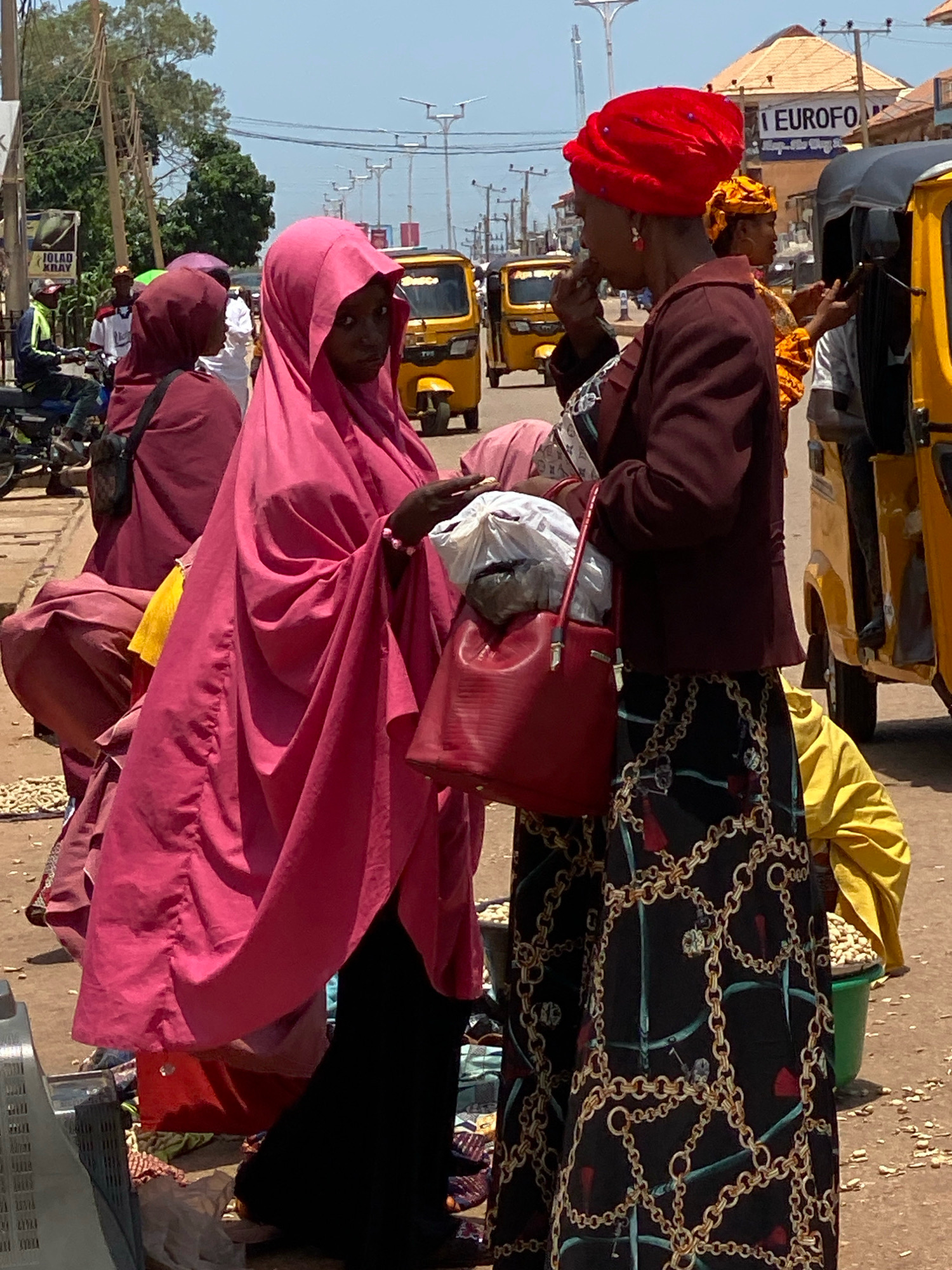 This image depicts a street scene in Sabon Tasha, Nigeria, on a bright, sunny day. The foreground is dominated by two women. The woman on the left is wearing a vibrant pink hijab and a matching, voluminous dress that covers her from head to toe. She is looking towards the right, her face partially obscured by the fabric. In her hands, she holds a white plastic bag filled with items, and a bright red handbag with a textured exterior is slung over her right forearm.
To her right stands another woman, taller and wearing a rich burgundy jacket over a black and gold patterned skirt. She has a striking red headscarf adorned with what appear to be small, sparkling embellishments, and she is looking downwards, her hand raised to her mouth, possibly eating or speaking. Her left arm is bent, holding the red handbag.
Behind these two women, the environment is bustling. Yellow auto-rickshaws, known as "Keke Napep," are visible on the street, suggesting local transportation. In the background, other individuals in traditional attire and motorcycles are present, hinting at a lively marketplace or a busy thoroughfare. Power lines crisscross the sky, and trees with green foliage can be seen. The ground is dusty and uneven, typical of many Nigerian streets. The presence of various goods, some in baskets, implies a commercial area. A sign with the text "EUROFOAM" is visible on a building in the background, indicating a commercial establishment.
