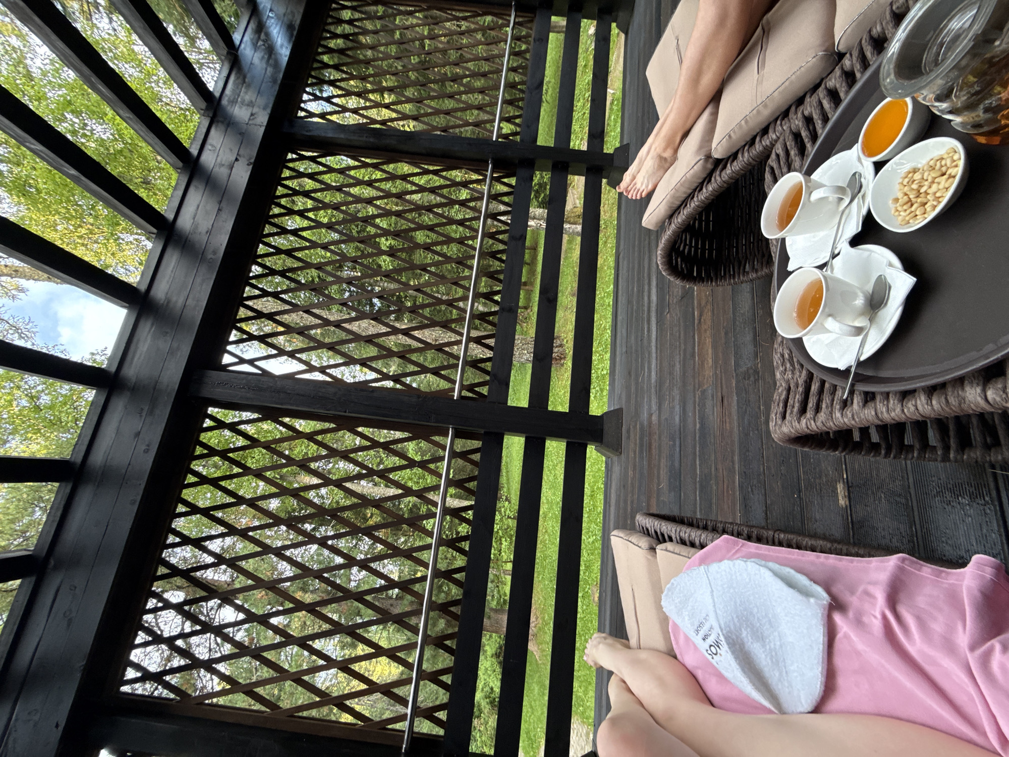 In a shaded outdoor seating area, possibly a resort or spa, two individuals are relaxing on lounge chairs. The structure appears to be a wooden pergola with dark wood beams and a diamond-patterned lattice screen. Through the screen, a lush green forest with tall trees and a bright sky with scattered clouds is visible.
In the foreground on the right, a person's legs are visible, resting on a brown woven lounge chair with light brown cushions. On a dark brown tray placed on another lounge chair in front of them are two white teacups with saucers and spoons, a small white oval dish filled with pine nuts, and an oval dish with orange preserves or jam. A glass carafe with an amber liquid, possibly tea or juice, is also on the tray.
In the foreground on the left, another person's legs are visible, resting on a similar lounge chair. A pink towel or blanket is draped over the chair, and a white folded towel with text is placed on top. The floor is made of dark wooden planks. The overall impression is one of relaxation and tranquility amidst a natural, forested environment. The text on the white towel might indicate a specific location or service.